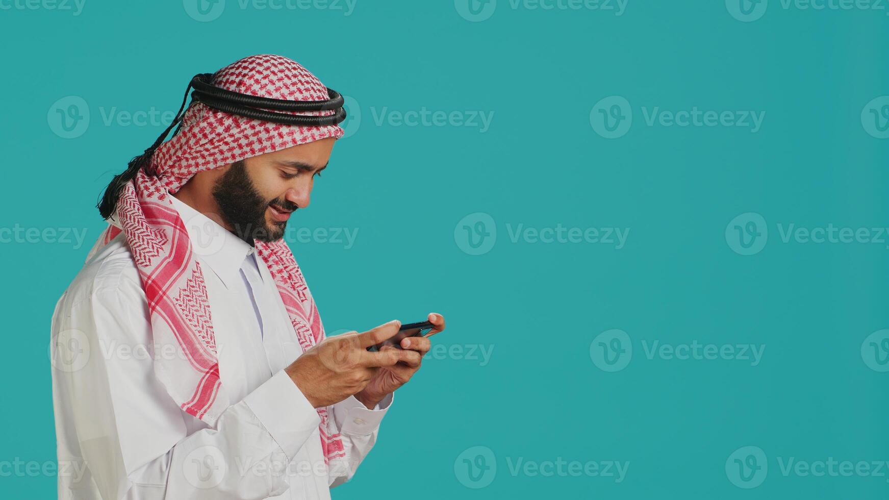 Arabic person playing mobile videogames on smartphone with tensed expression, wears islamic headscarf and thobe. Muslim gamer enjoying online gaming experience on phone app in studio. photo