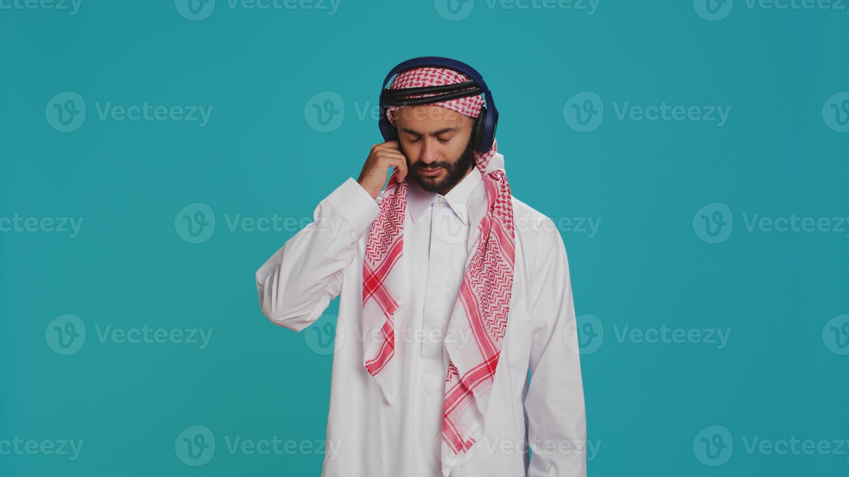 Joyful person dancing on songs in studio, listening to modern music on headphones and having fun on camera. Middle eastern adult in traditional clothing showing groovy dance moves. photo