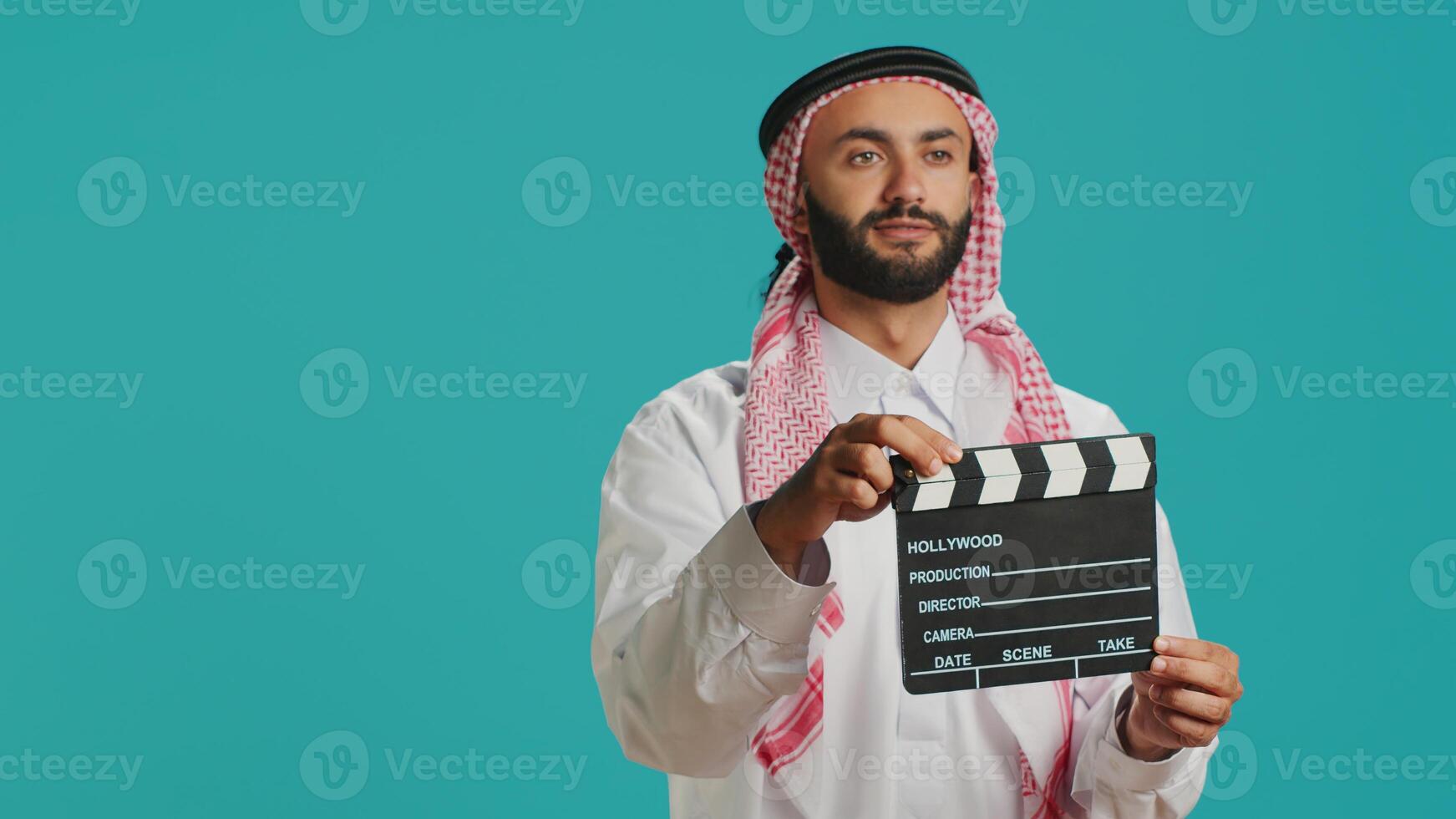 Middle eastern guy with clapboard used for scene takes on movie industry, young director using cinematography filming slate. Arab filmmaker wearing traditional muslim attire and scarf. photo