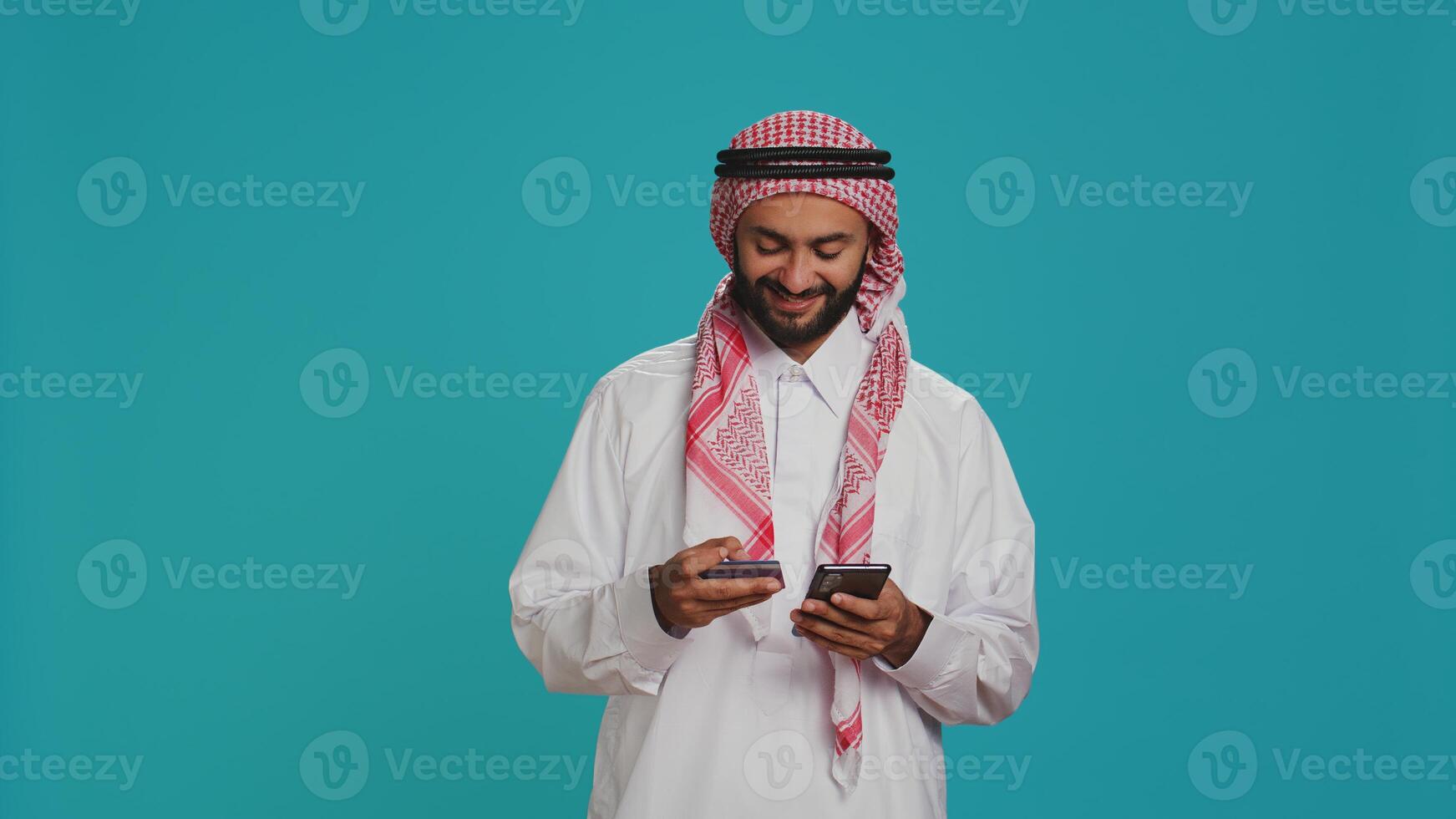 Joyful model using card to make payment while shopping on clothing store website using smartphone app. Middle eastern adult in islamic costume doing banking transfer to buy on sale. photo