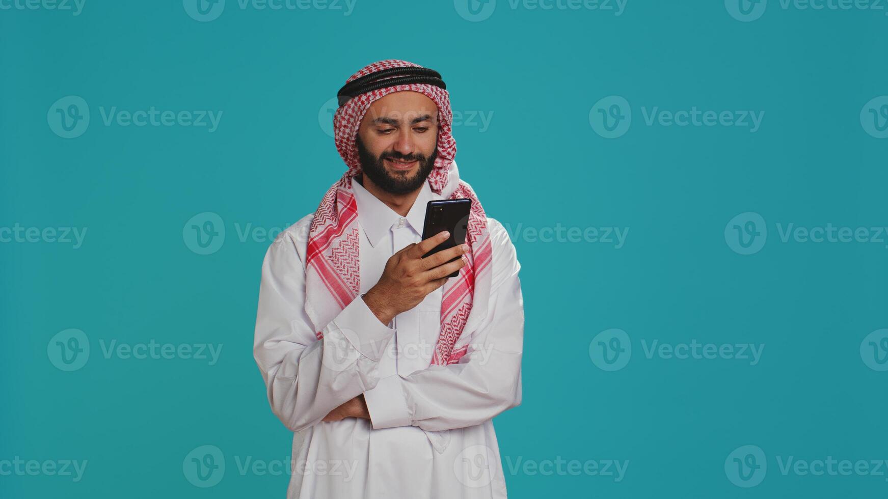 Muslim man in traditional clothing using smartphone, engaged in online social media conversation on camera. Middle eastern guy wearing arabic headscarf and gown, navigating web. photo