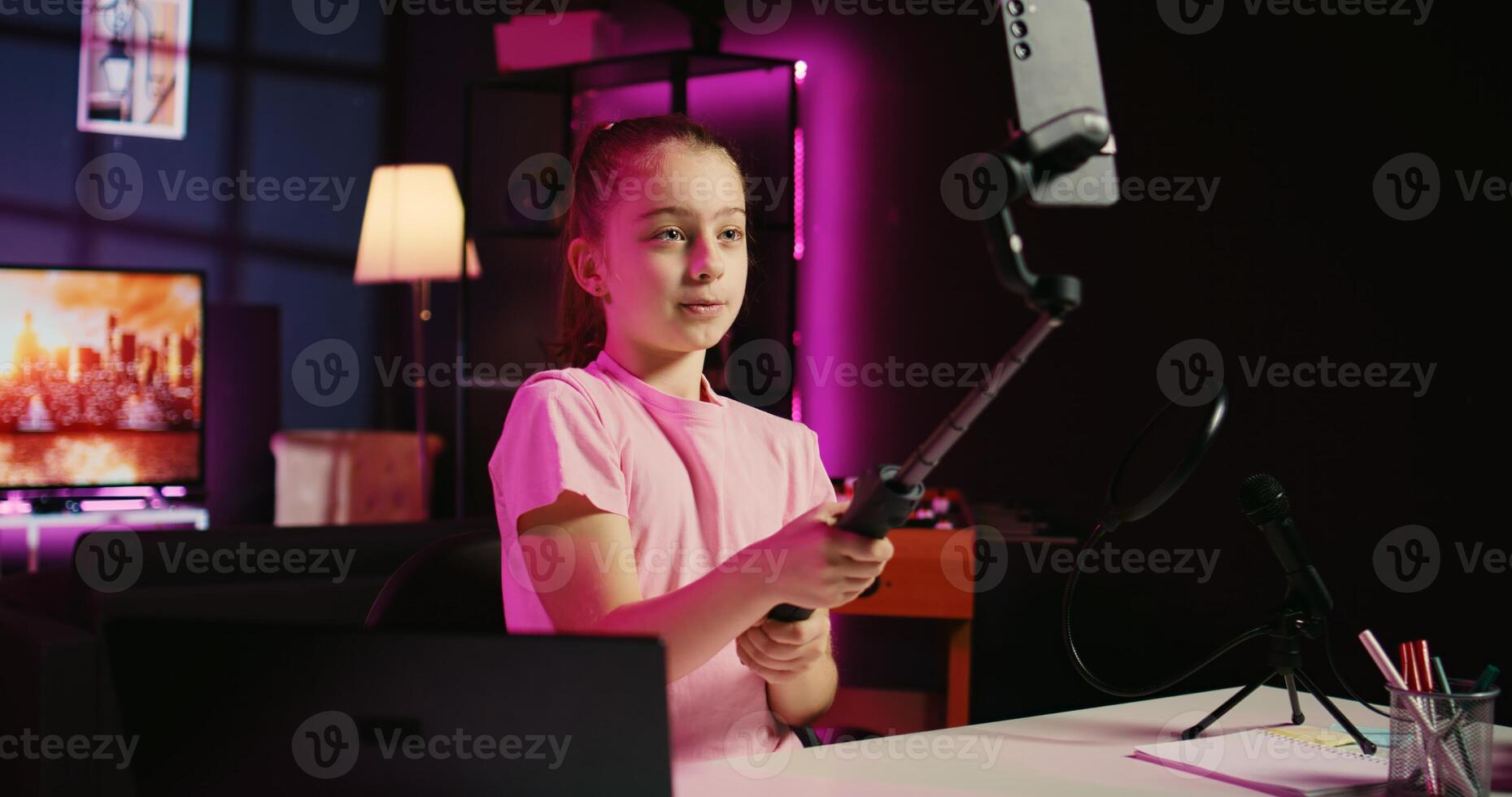 Child filming with smartphone attached to selfie stick in living room used as professional studio for recording vlogs. Happy kid captures footage with phone in neon lit apartment, discussing topics photo