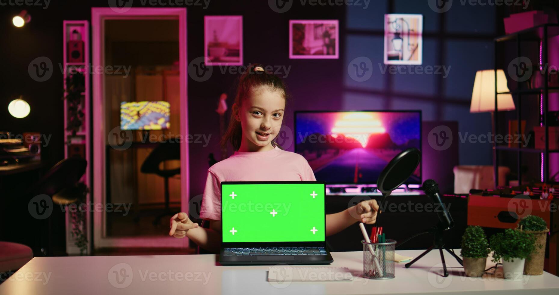 Cheerful child in home studio presenting advertisement from sponsoring brand on green screen laptop. Gen Z girl using chroma key device to present promotion from media partner to followers photo