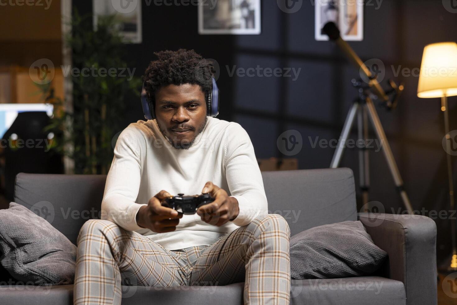 Gamer using headphones mic to discuss with teammates while playing games and enjoy leisure time. BIPOC man in apartment entertained by videogame on console, using gamepad photo