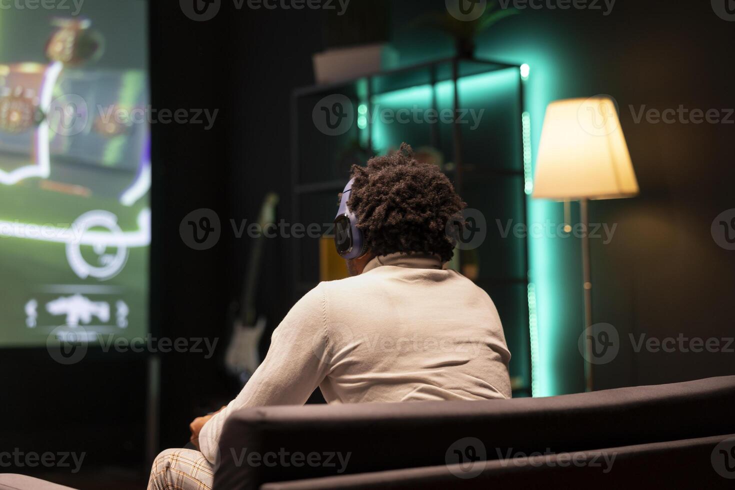 BIPOC man playing SF FPS videogame on widescreen smart TV, having fun in neon lit apartment. Gamer competing in online multiplayer game, using headphones to talk with teammates photo