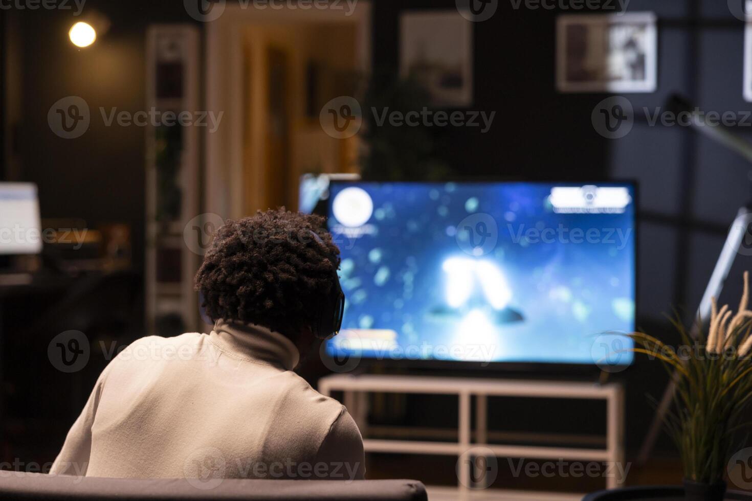 Gamer at home gaming and enjoying leisure time using television set. African american man sitting on couch in living room, playing videogame on smart TV to take off steam photo