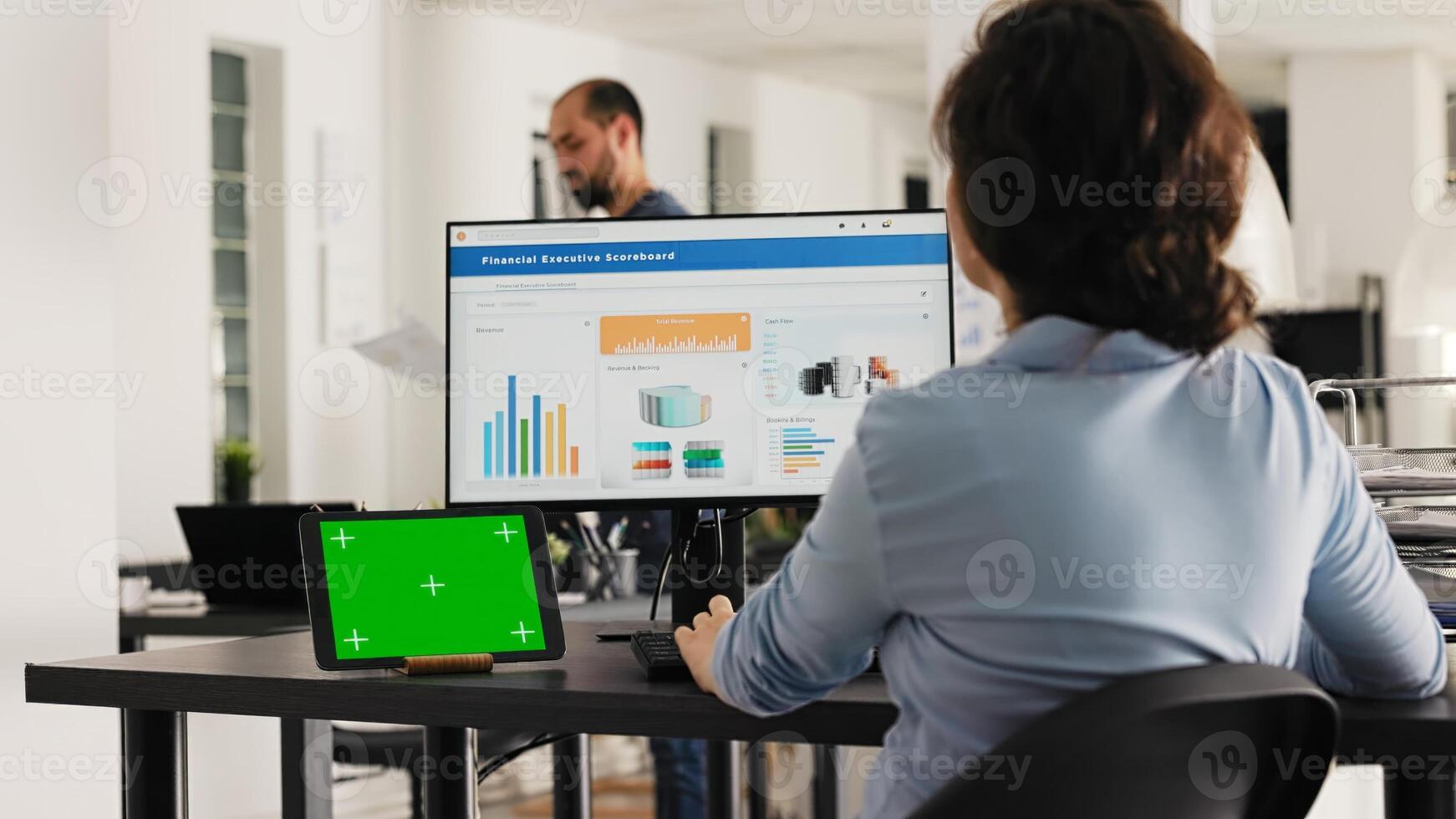 Analyst working at office desk with greenscreen shown on tablet, looking at blank copyspace layout on device while she checks sales data. Employee uses chromakey template. Handheld shot. photo