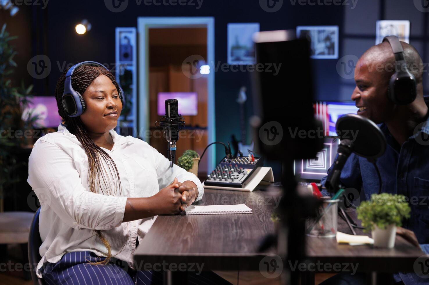 Influential African American host engages with guest, using quality podcast equipment in their stylish home studio. Black couple utilizing smartphone on tripod to record their online talk show. photo