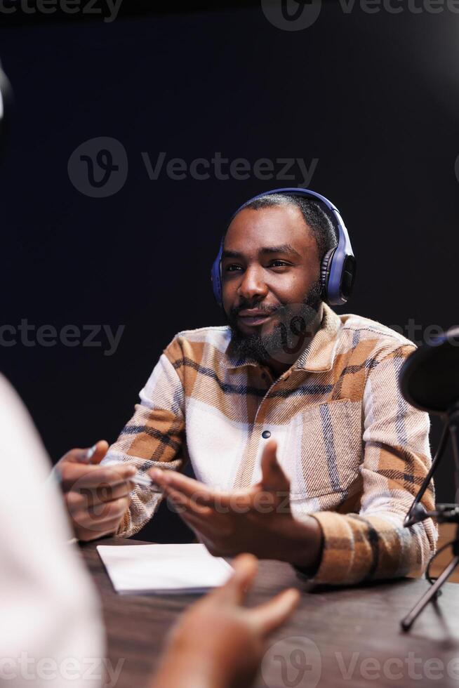African American man creates internet radio programs at home with audio gadgets. Black male content creator using wireless headphones, a microphone, and podcast equipment while talking with guest. photo