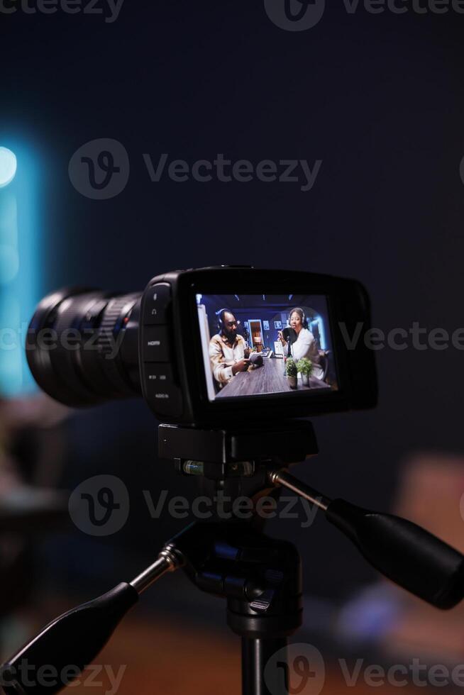 Detailed view of professional camera recording male journalist with notebook interviewing famous blogger. Selective focus of digital device on tripod filming black couple doing live talk show. photo