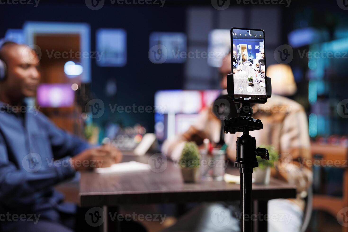 Smartphone is focused selectively on a tripod while recording two African American bloggers discussing various issues. Black male influencers gather in a home studio to film a show on a mobile device. photo