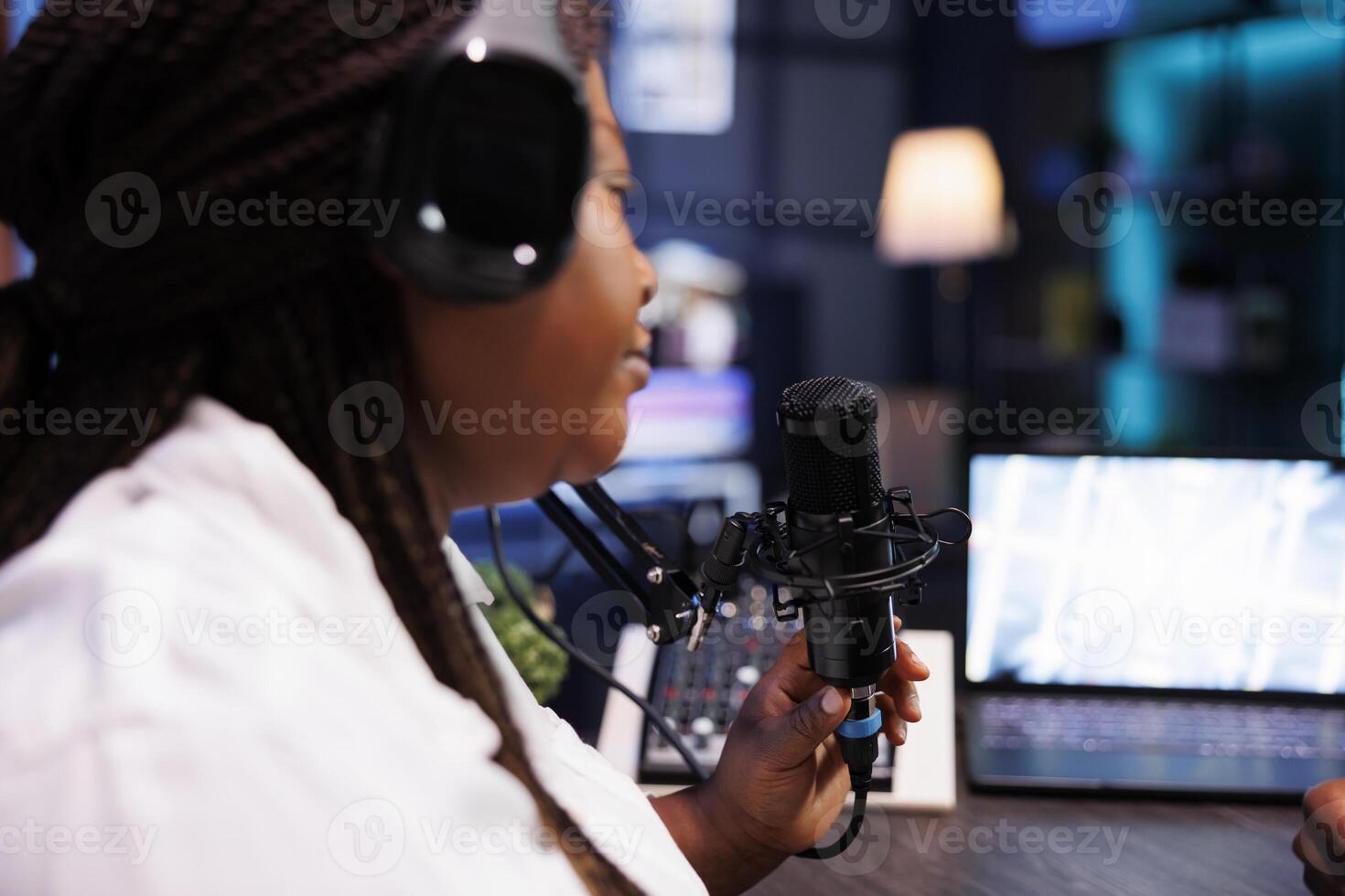 Black female talk show host speaking with online listeners during podcast episode, wearing wireless headphones and talking into microphone. Influencer utilizing audio equipment in home studio. photo