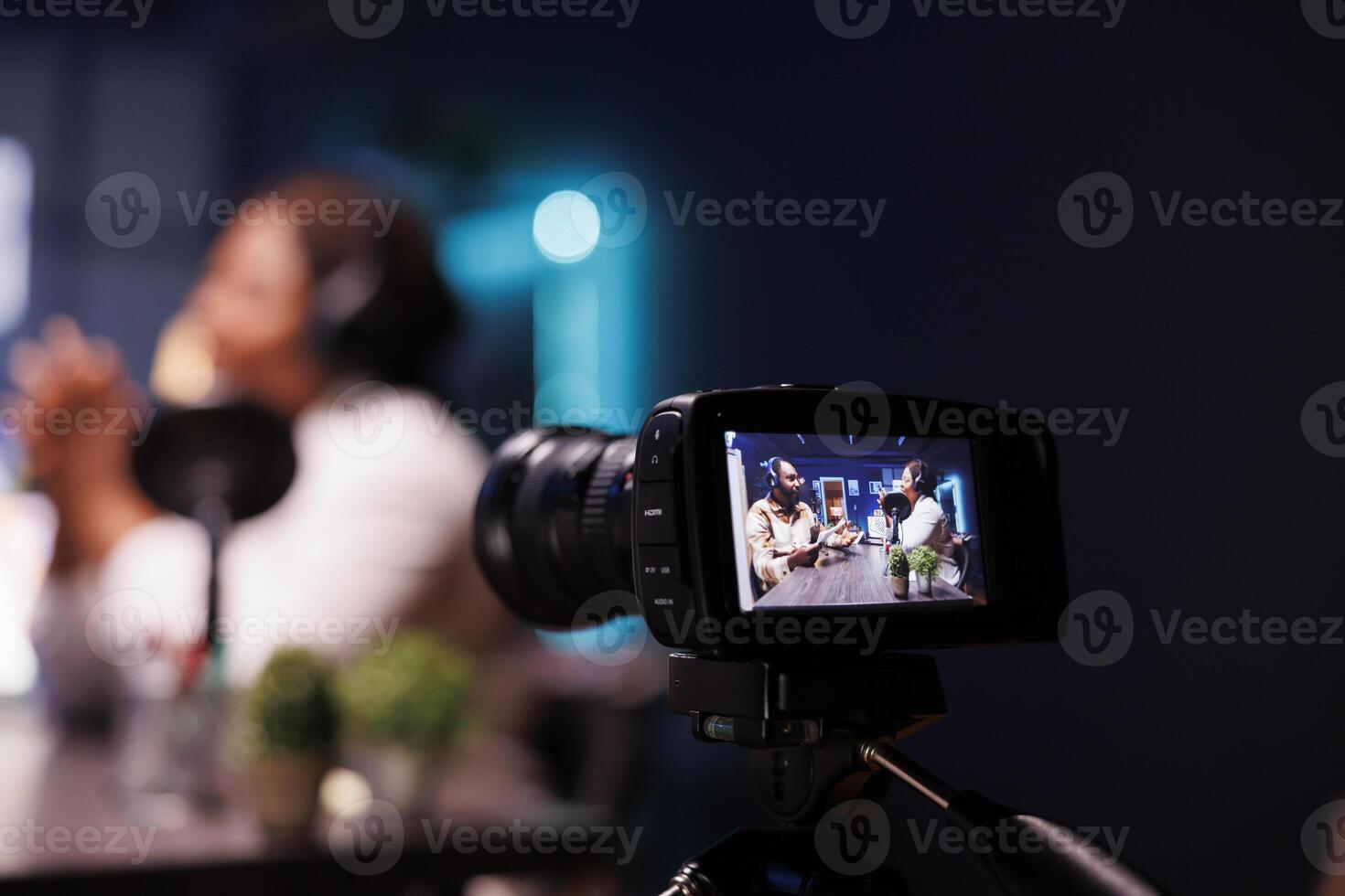 Closeup of camera in front of black couple recording an online show. African male and female vloggers speaking into microphone during a live stream, discussing in podcast while wearing headphones. photo