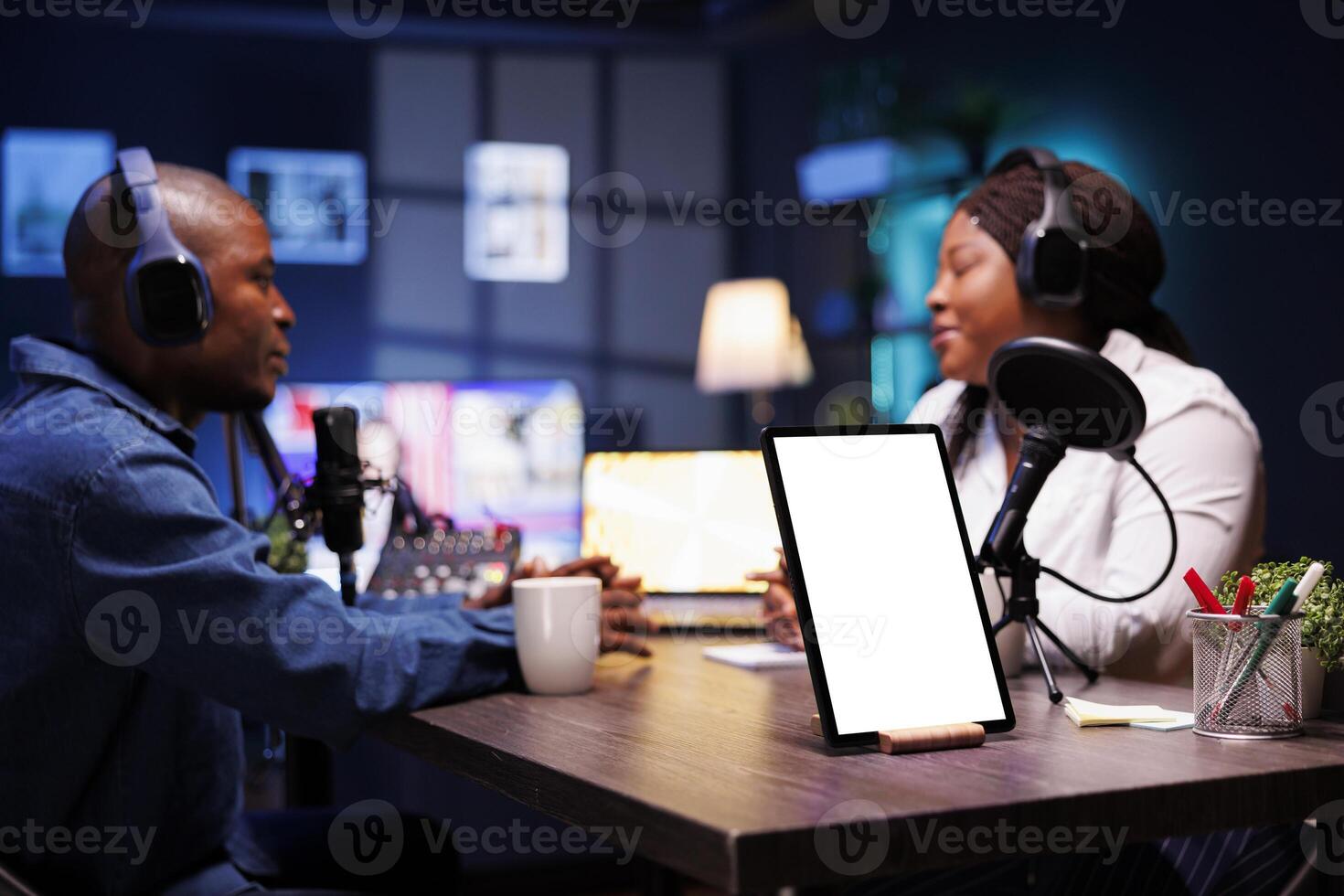 Two African American vloggers wearing headphones are recording a live podcast while a tablet on desk shows a white screen with mockup template. Digital device on table displays isolated chromakey. photo