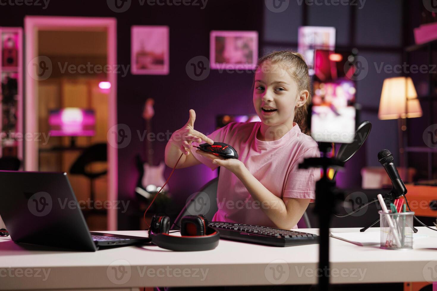 Cute girl in studio reviewing neon lit gaming mouse in front of camera in personal studio. Joyous kid filming electronics haul, presenting Bluetooth wireless computer peripheral to viewers photo