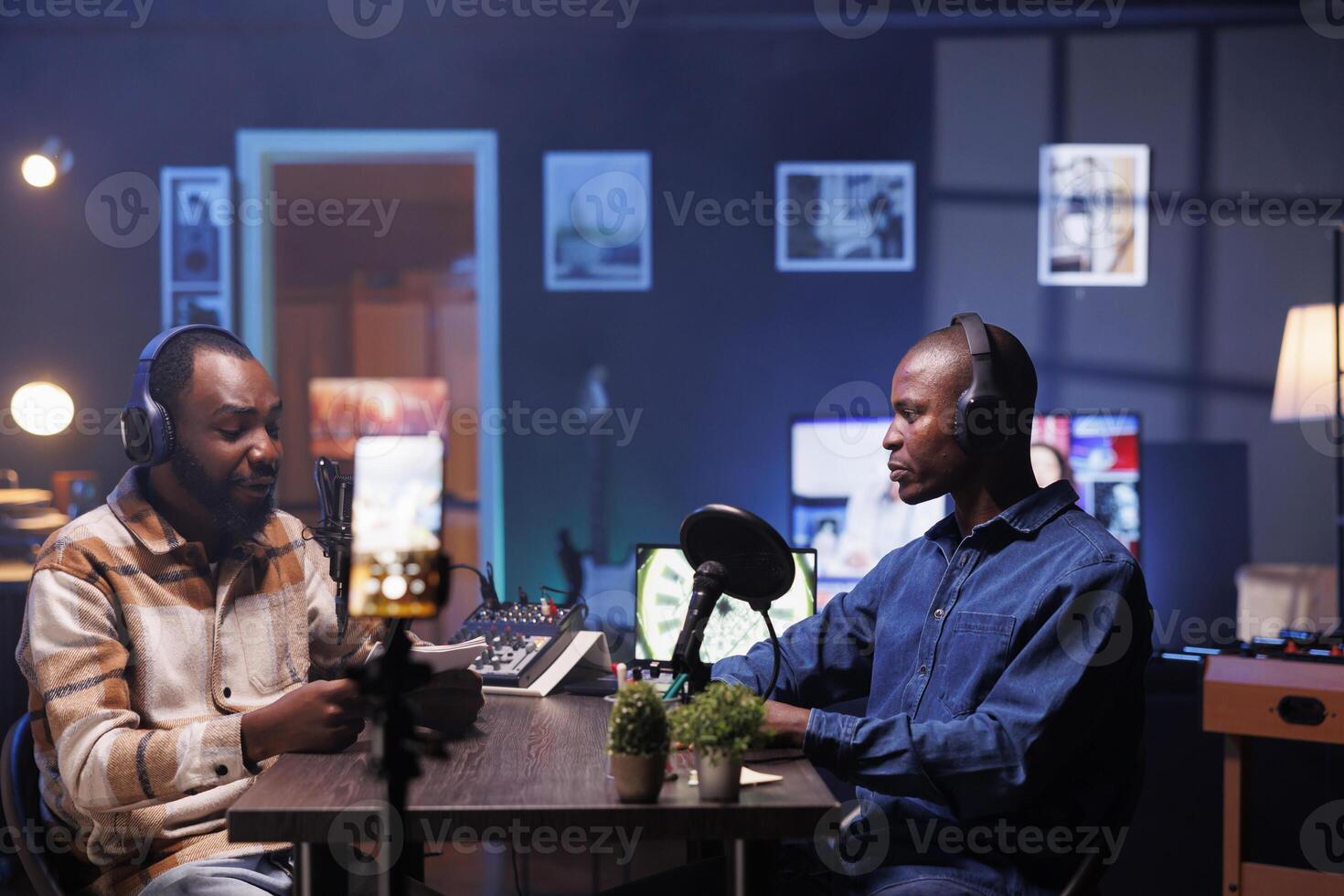Vlogger is recording an interview with smartphone on tripod in studio while reviewing notes to ask quality questions to guest. Journalist using mobile device to film vlog while talking on mic with man photo