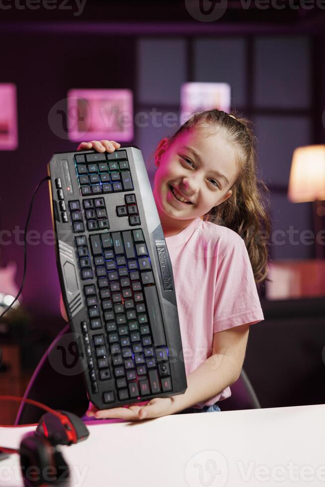 Happy gen Z content creator filming keyboard review in home studio, showing typing capabilities. Smiling small girl showcasing RGB illuminated computer peripheral to audience photo