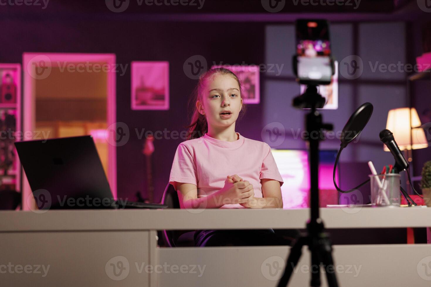 Child doing content creation, using cellphone on tripod stabilizer to film vlog channel intro. Young online star greets children audience, presenting today topic, recording with mobile phone photo
