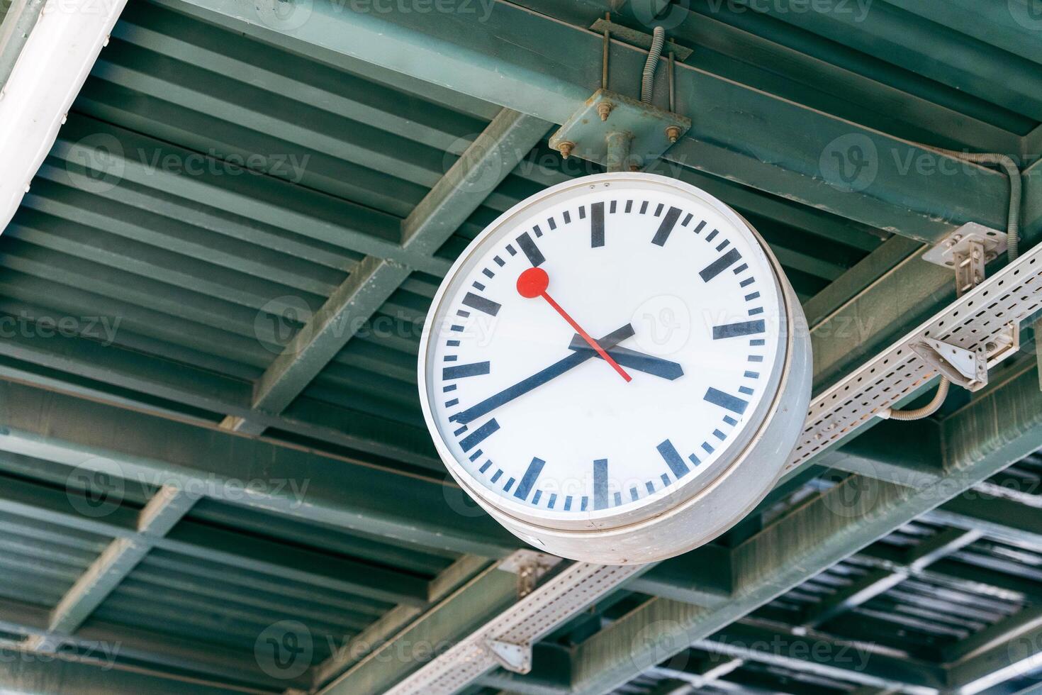 A large clock hanging from a metal ceiling under a train station canopy photo