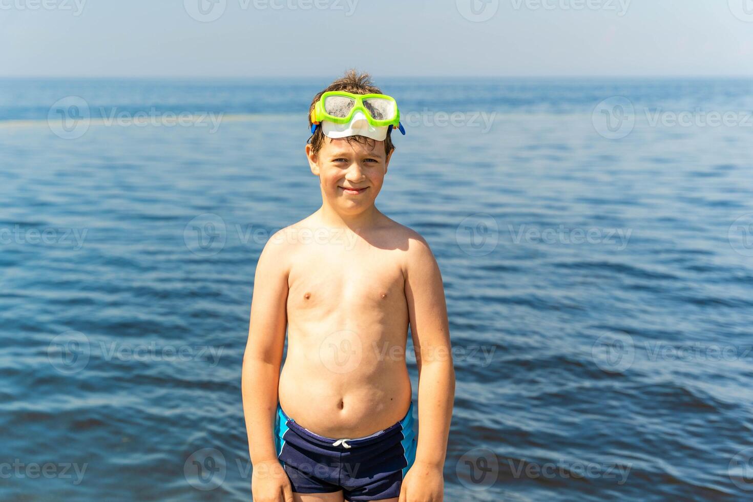 Young boy in swim goggles and trunks stands by water at beach photo
