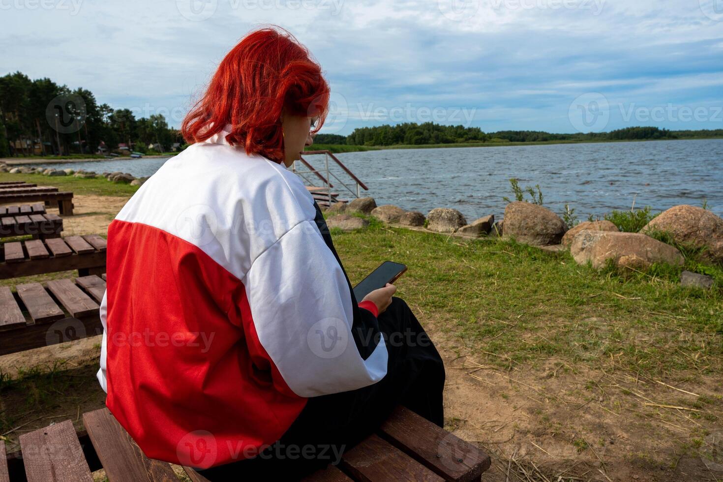 Stylish Generation Z Redhead Girl Sitting by Quiet Lake Using Smartphone photo