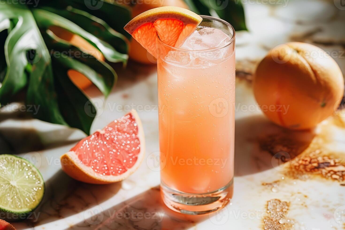 A highball glass filled with a refreshing grapefruit iced beverage, garnished with a grapefruit wedge. Surrounding are fresh grapefruits, lime, and monstera leaves, all set on a marble surface. photo