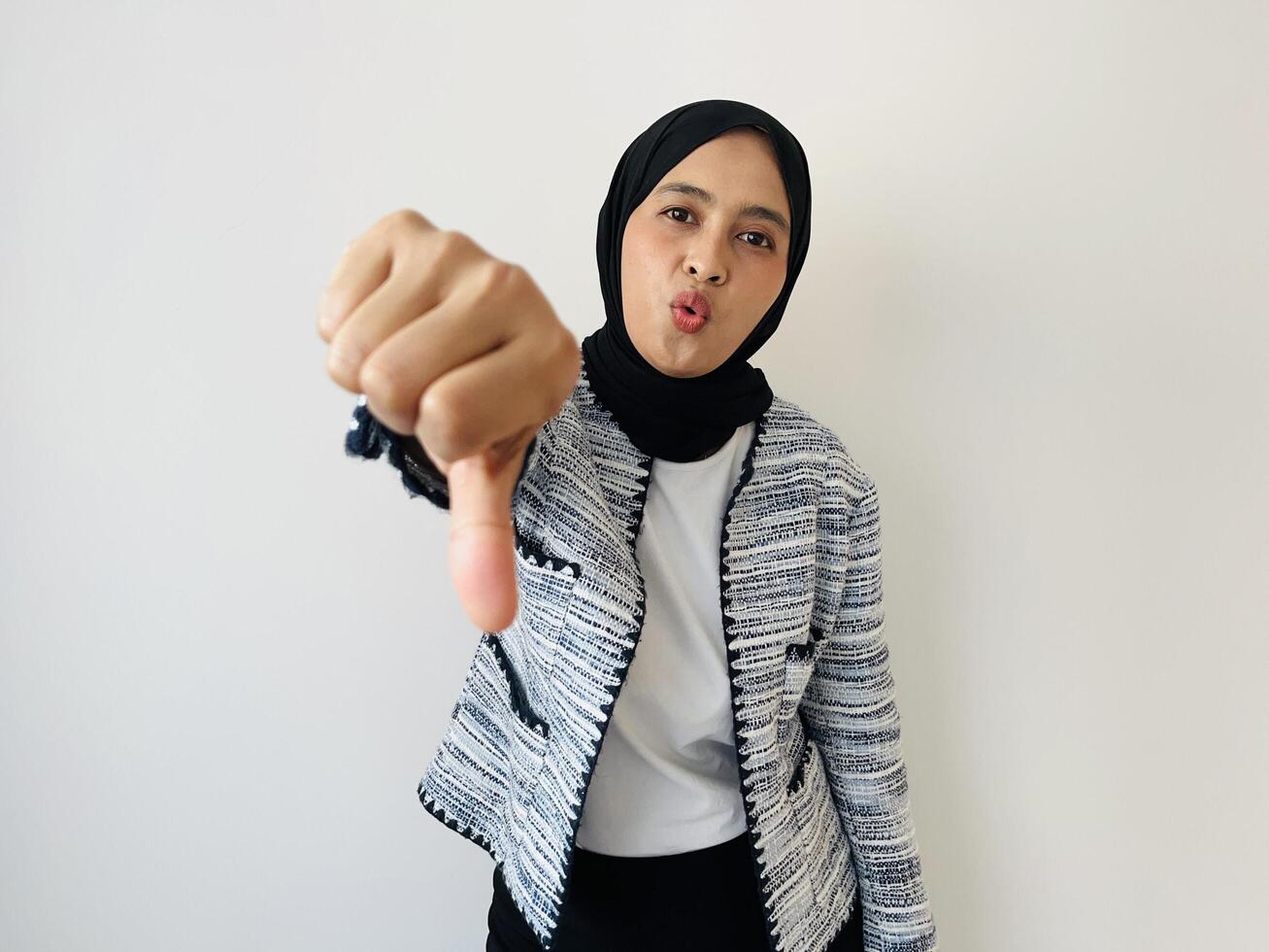 Dislike. Upset Asian woman wearing black hijab and tweed blazer gives thumbs down sign gesture, expressing discontent, disapproval, dissatisfied bad work, mistake isolated by white background photo
