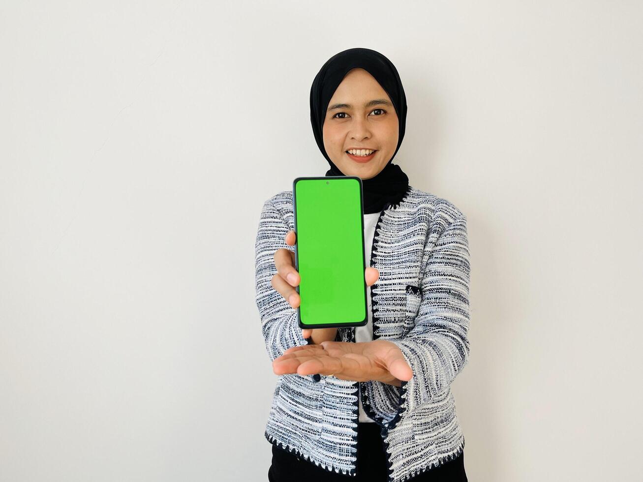 A smiling young Asian Muslim woman show smartphone green color of the screen with positive expression, dressed in tweed blazer and black hijab feeling happy isolated by white background. Mobile phone photo
