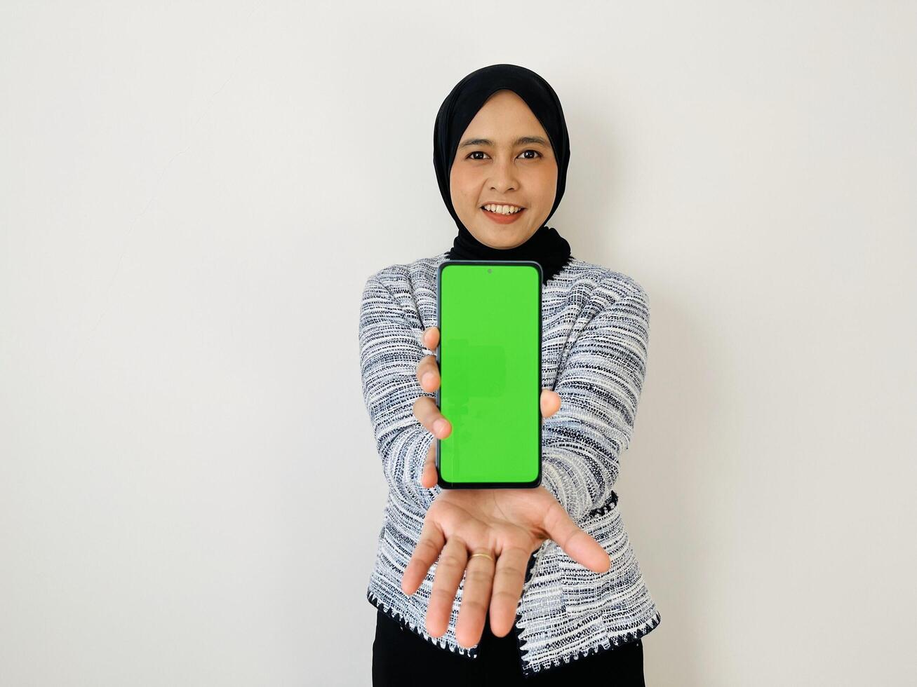 A smiling young Asian Muslim woman show smartphone green color of the screen with positive expression, dressed in tweed blazer and black hijab feeling happy isolated by white background. Mobile phone photo