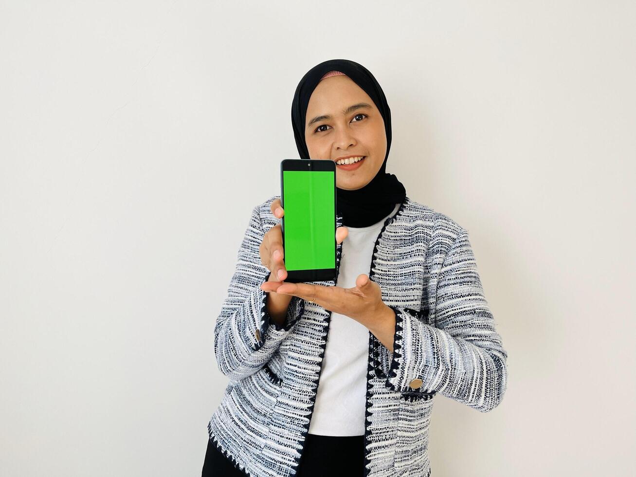 A smiling young Asian Muslim woman show smartphone green color of the screen with positive expression, dressed in tweed blazer and black hijab feeling happy isolated by white background. Mobile phone photo