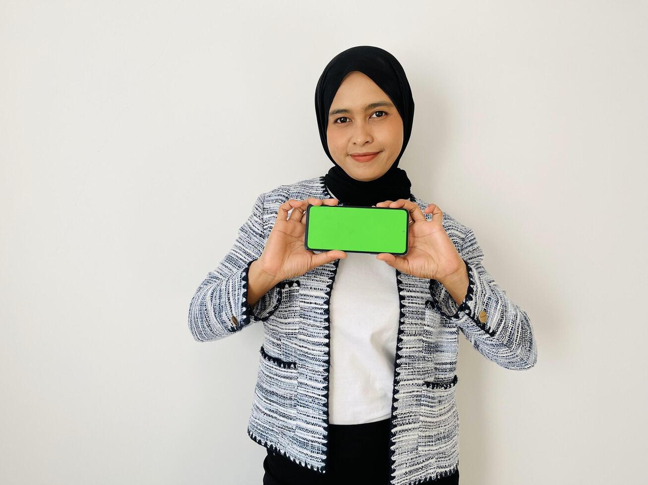 A smiling young Asian Muslim woman show smartphone green color of the screen with positive expression, dressed in tweed blazer and black hijab feeling happy isolated by white background. Mobile phone photo
