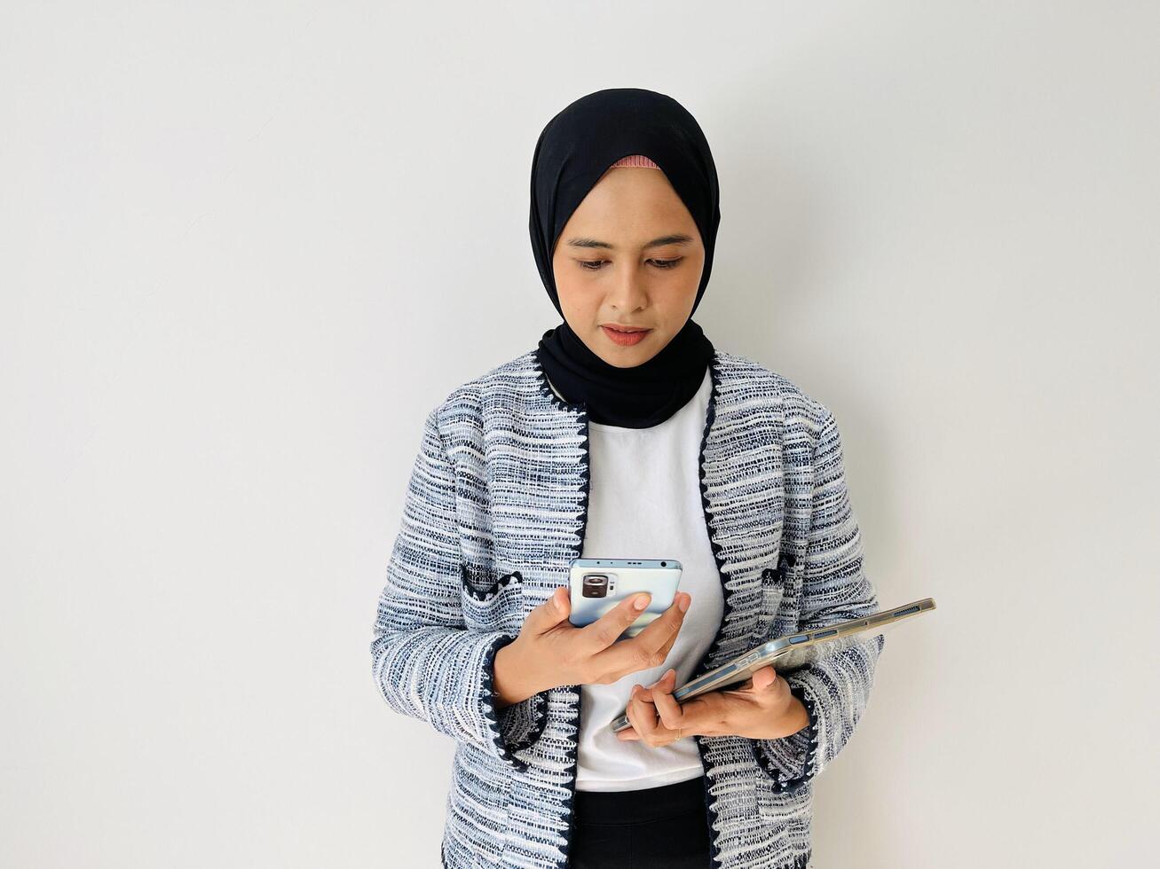 Young Asian woman smiling while using mobile phone. Standing female wearing black hijab and tweed blazer isolated on white background. photo