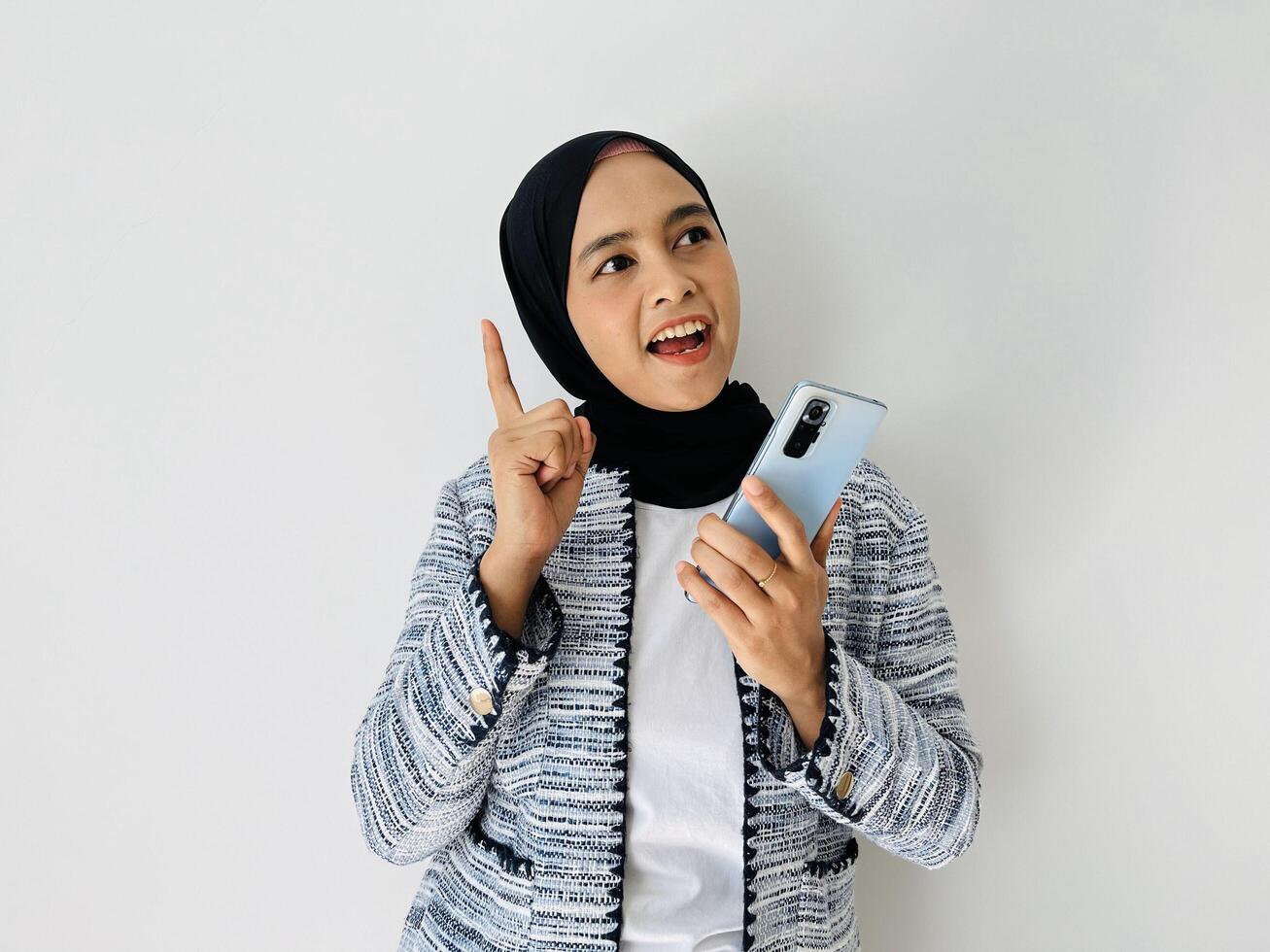 Portrait of happy young Muslim Asian woman got good ideas and looking up while holding phone. Got solution. Excited smart thoughtful, pointing up, eureka moment white background. photo