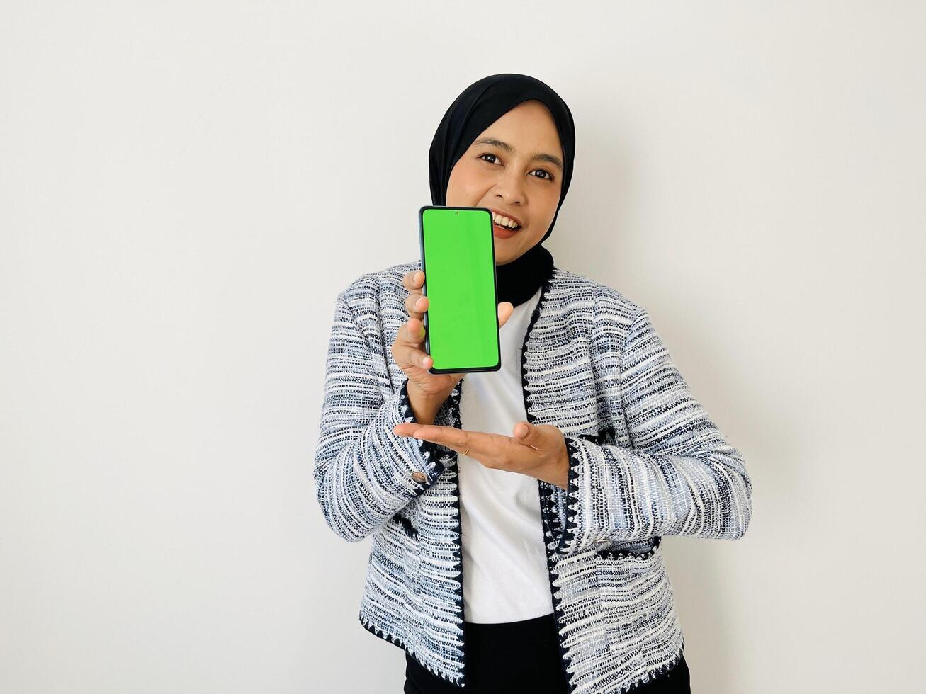 A smiling young Asian Muslim woman show smartphone green color of the screen with positive expression, dressed in tweed blazer and black hijab feeling happy isolated by white background. Mobile phone photo