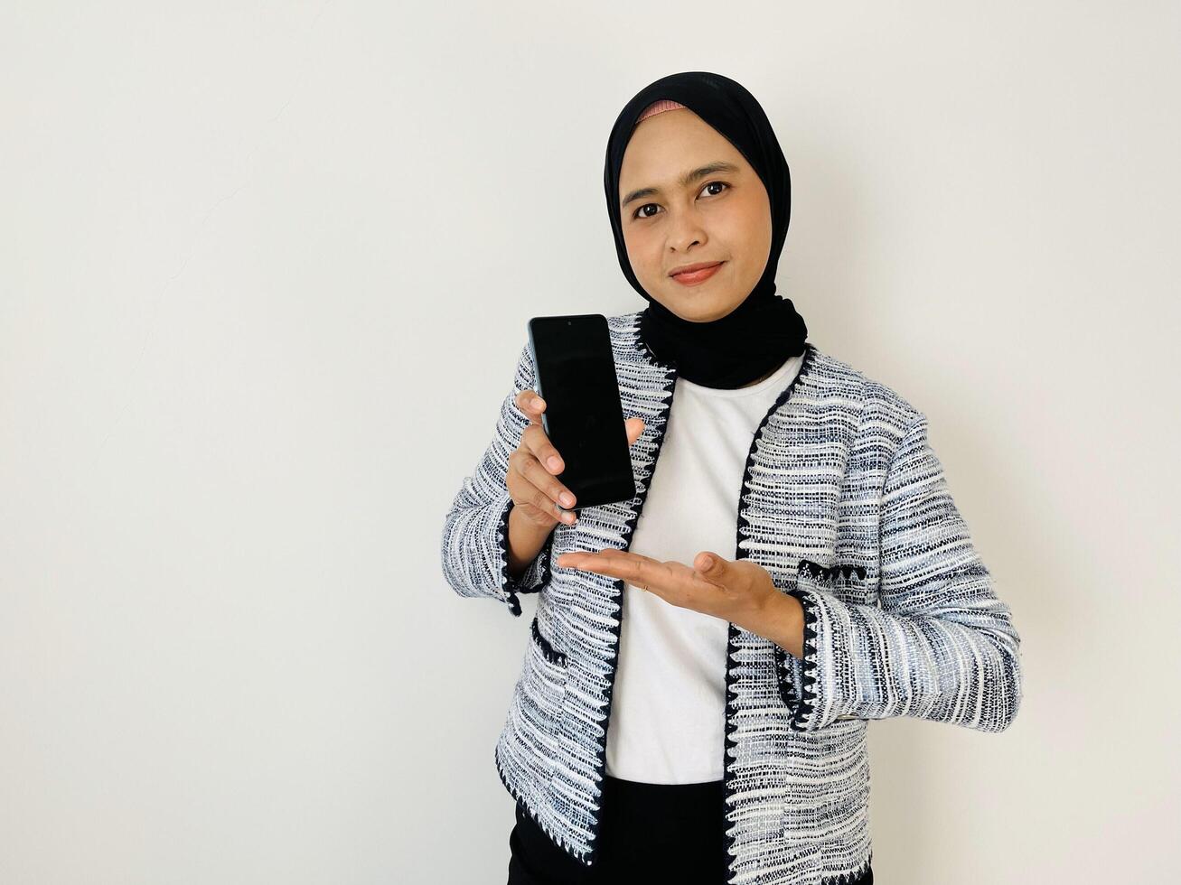 A smiling young Asian Muslim woman show smartphone screen with positive expression, dressed in tweed blazer and black hijab feeling happy isolated by white background photo