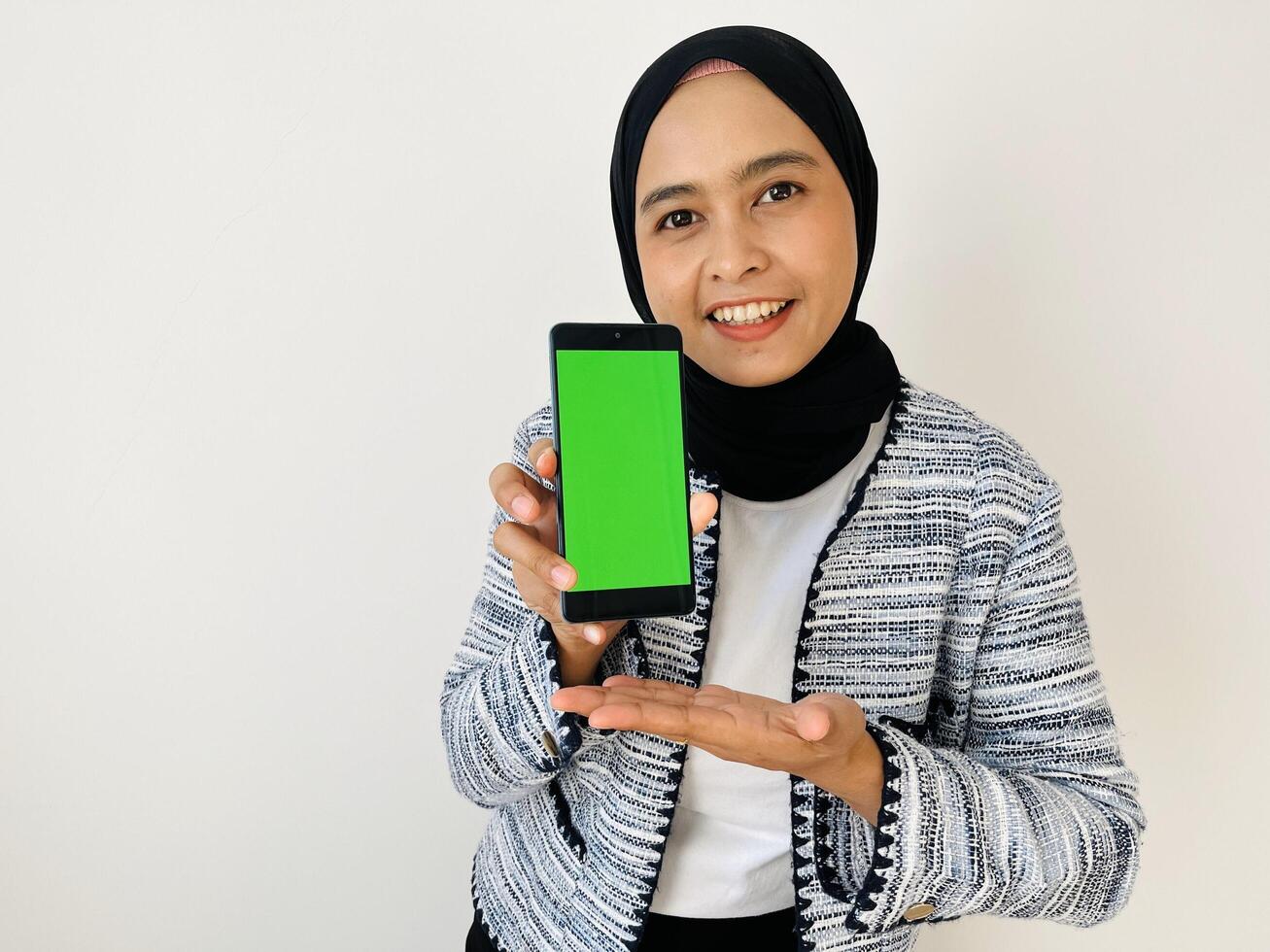 A smiling young Asian Muslim woman show smartphone green color of the screen with positive expression, dressed in tweed blazer and black hijab feeling happy isolated by white background. Mobile phone photo