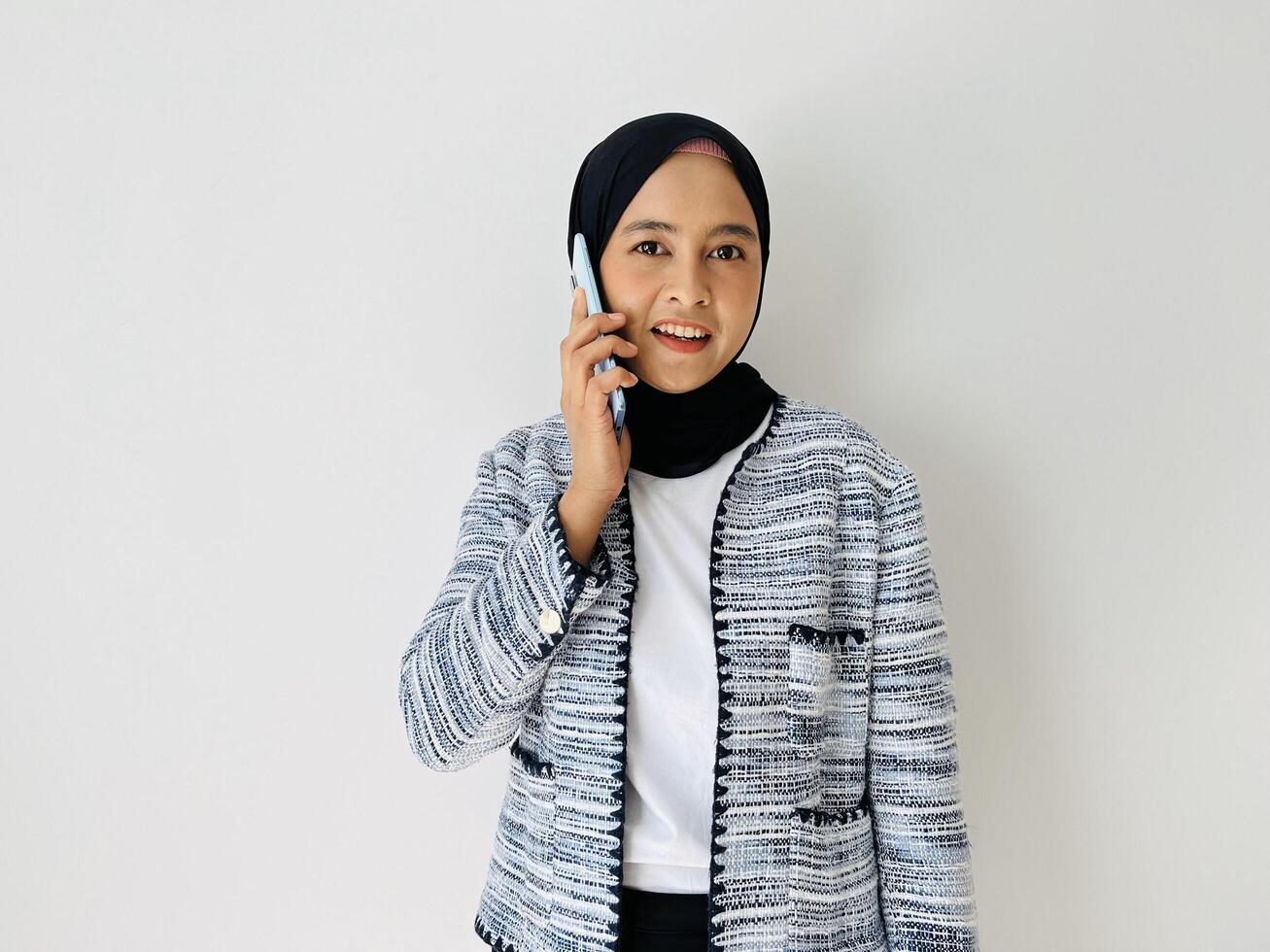 Young Asian woman smiling while using mobile phone having call. Standing female wearing black hijab and tweed blazer isolated on white background. photo