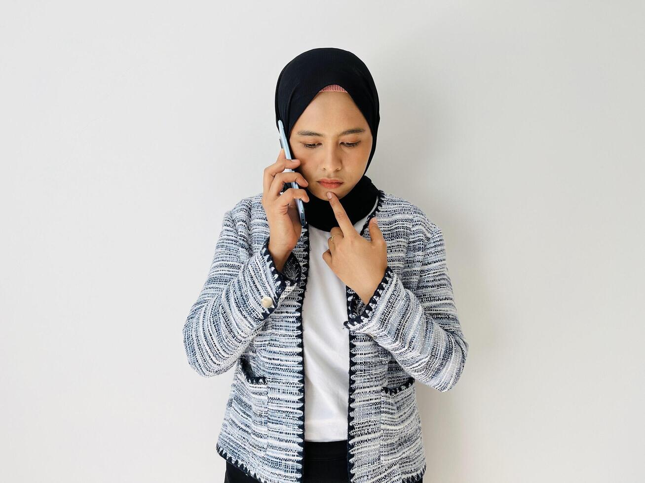 Young Asian woman using mobile phone having doubts while looking down. Female wearing wearing black hijab and tweed blazer isolated on white background. photo