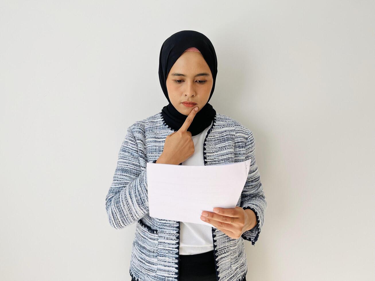 Young Asian woman thinking while reading document. Standing read report, planning focusing on contract. Female professional wearing black hijab and tweed blazer isolated by white background. photo