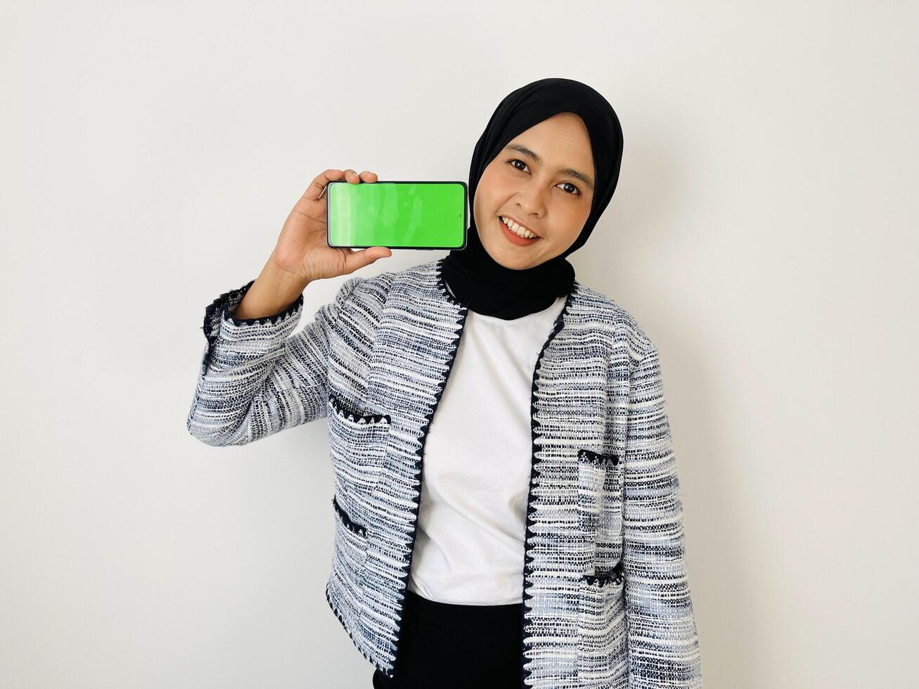 A smiling young Asian Muslim woman show smartphone green color of the screen with positive expression, dressed in tweed blazer and black hijab feeling happy isolated by white background. Mobile phone photo