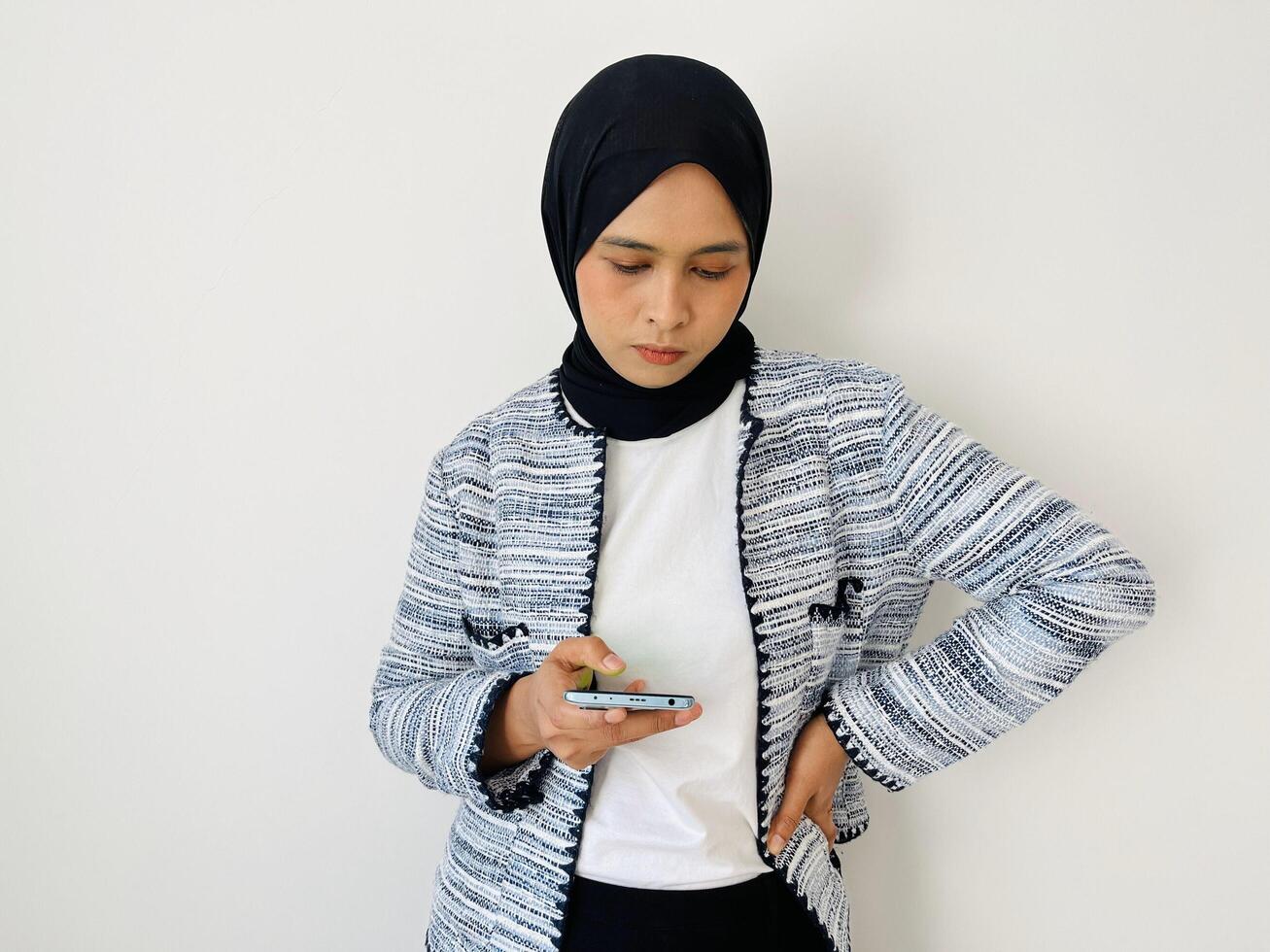 A young Asian Muslim woman holds and looks at a smartphone in annoyance with her other hand on her waist photo