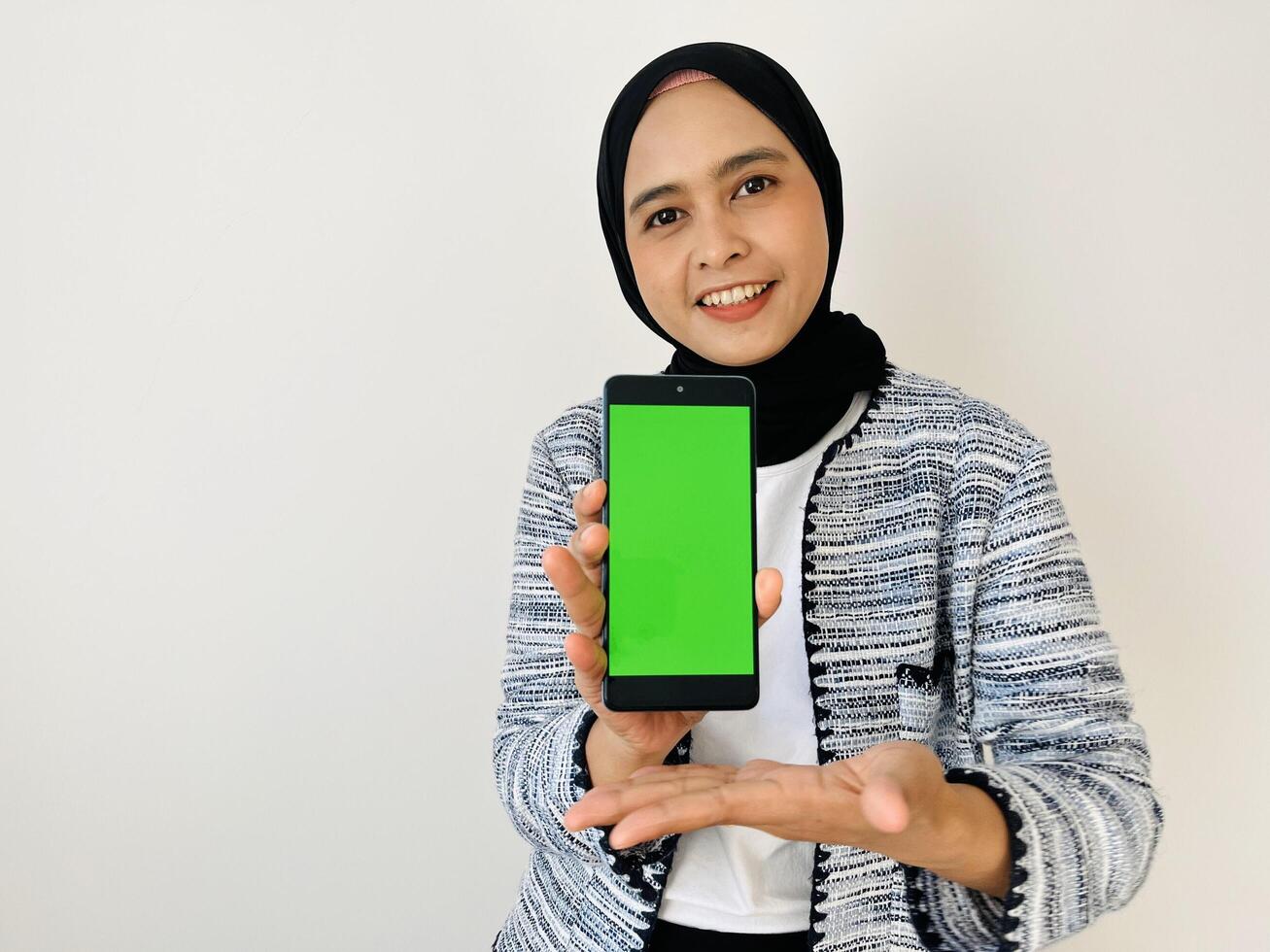 A smiling young Asian Muslim woman show smartphone green color of the screen with positive expression, dressed in tweed blazer and black hijab feeling happy isolated by white background. Mobile phone photo