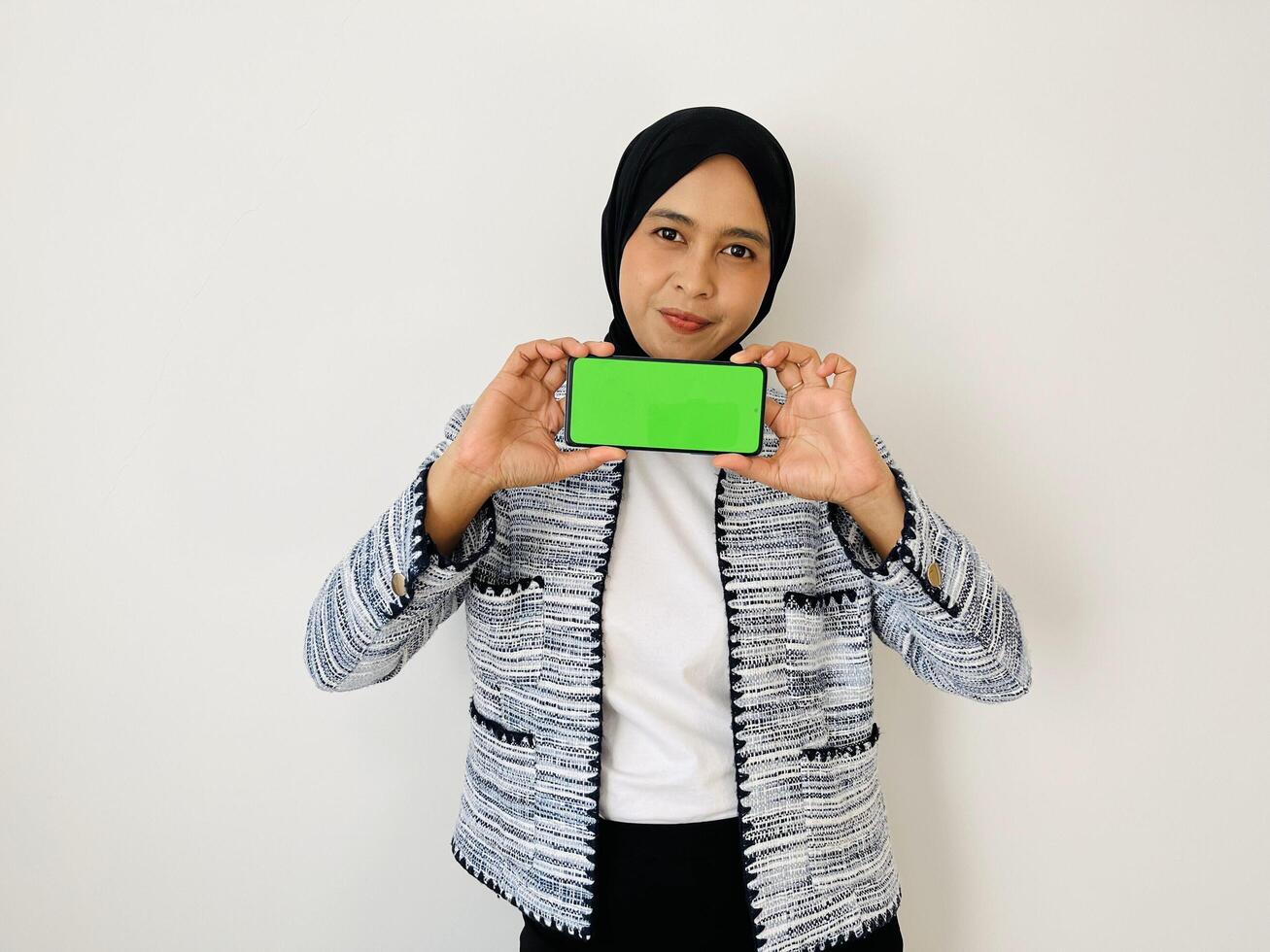 A smiling young Asian Muslim woman show smartphone green color of the screen with positive expression, dressed in tweed blazer and black hijab feeling happy isolated by white background. Mobile phone photo