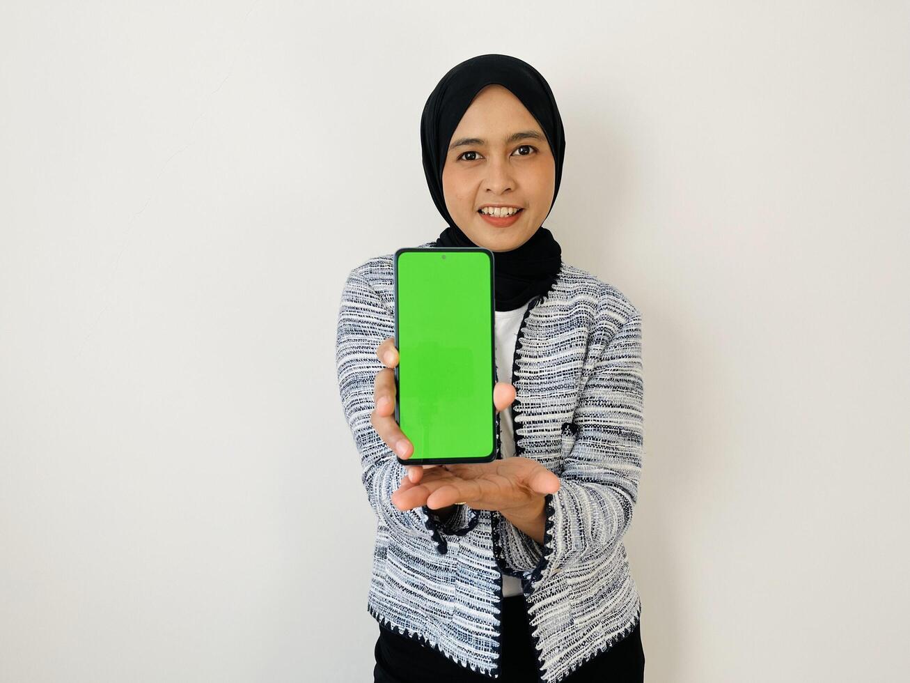 A smiling young Asian Muslim woman show smartphone green color of the screen with positive expression, dressed in tweed blazer and black hijab feeling happy isolated by white background. Mobile phone photo