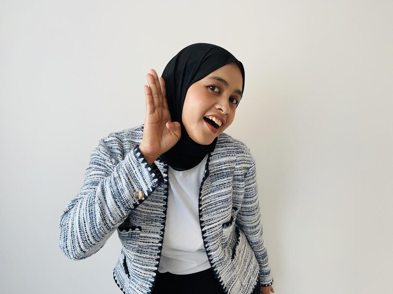 Amazed beautiful Muslim Asian woman puts a hand to the ear to hear better. Young woman listening something over white background. Deafness concept. Woman holds her hand near ear and listens carefully photo