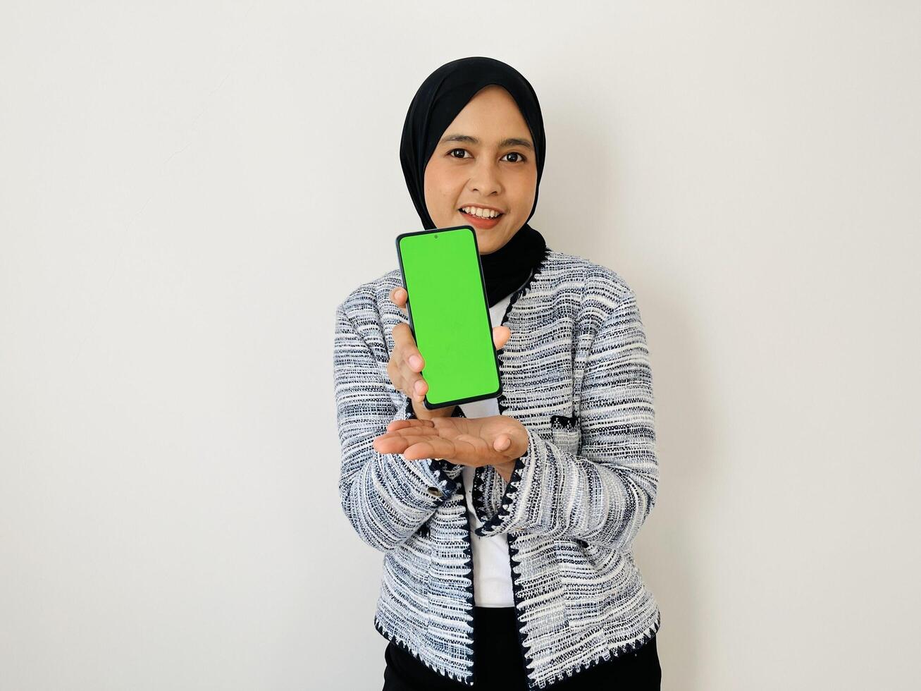 A smiling young Asian Muslim woman show smartphone green color of the screen with positive expression, dressed in tweed blazer and black hijab feeling happy isolated by white background. Mobile phone photo