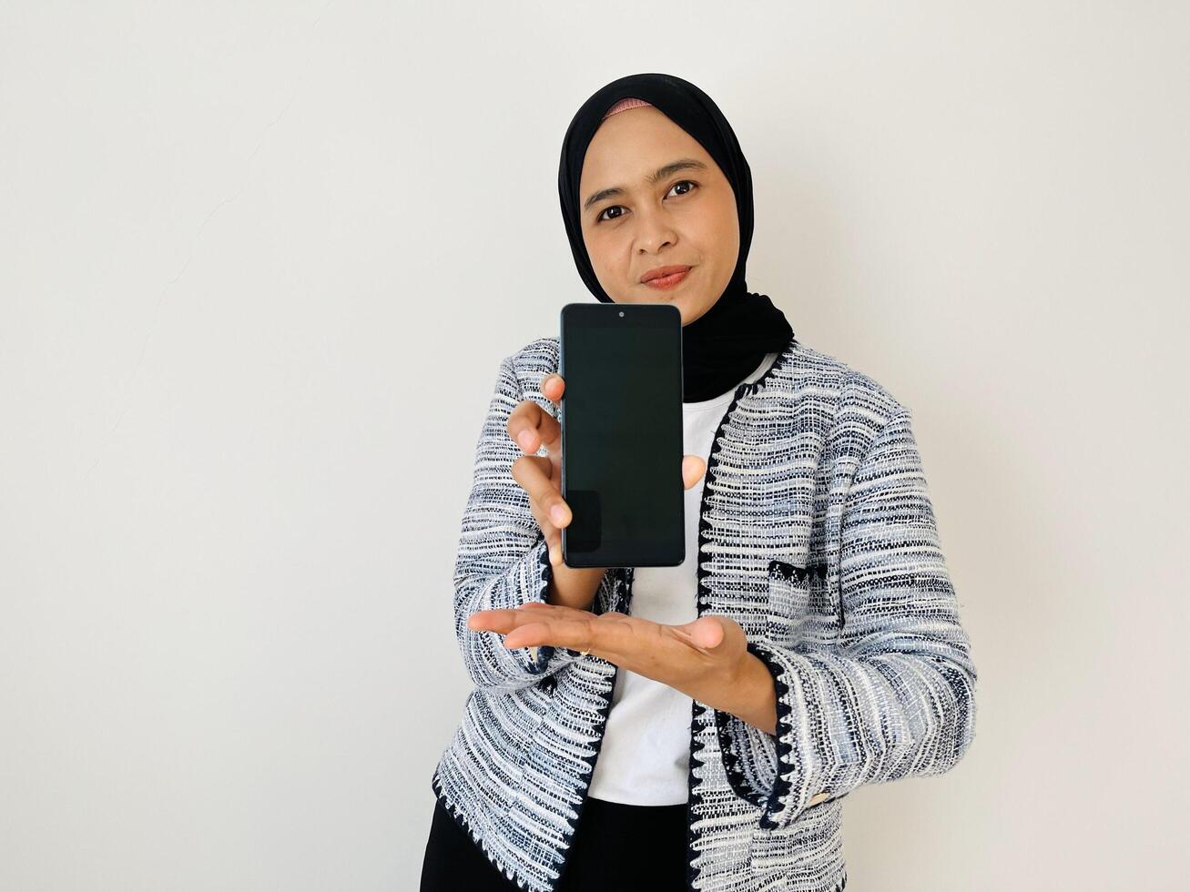 A smiling young Asian Muslim woman show smartphone screen with positive expression, dressed in tweed blazer and black hijab feeling happy isolated by white background photo
