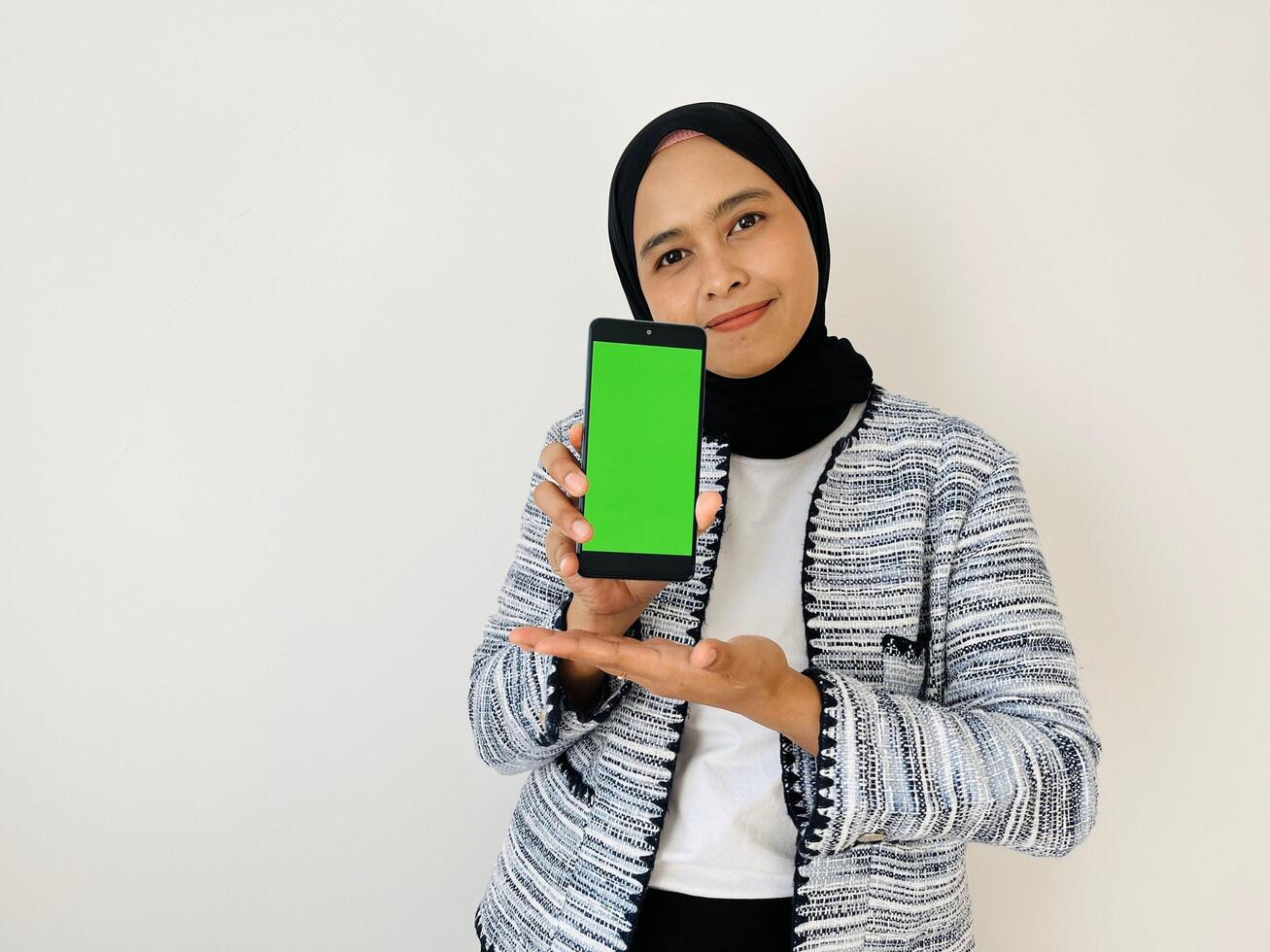 A smiling young Asian Muslim woman show smartphone green color of the screen with positive expression, dressed in tweed blazer and black hijab feeling happy isolated by white background. Mobile phone photo