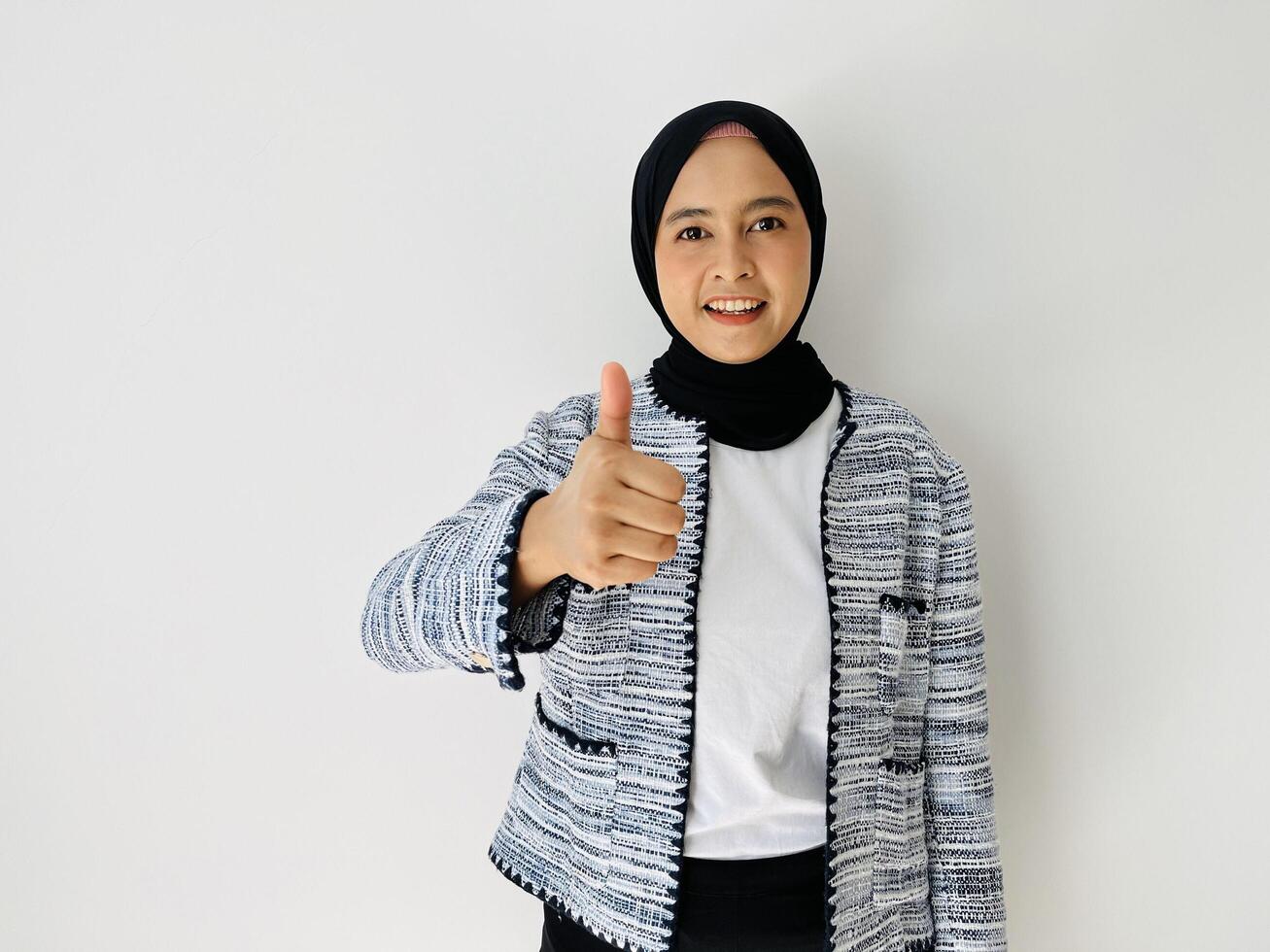 Smiling Asian woman wearing black hijab and tweed blazer gives thumbs up hand gesture of approval isolated by white background photo