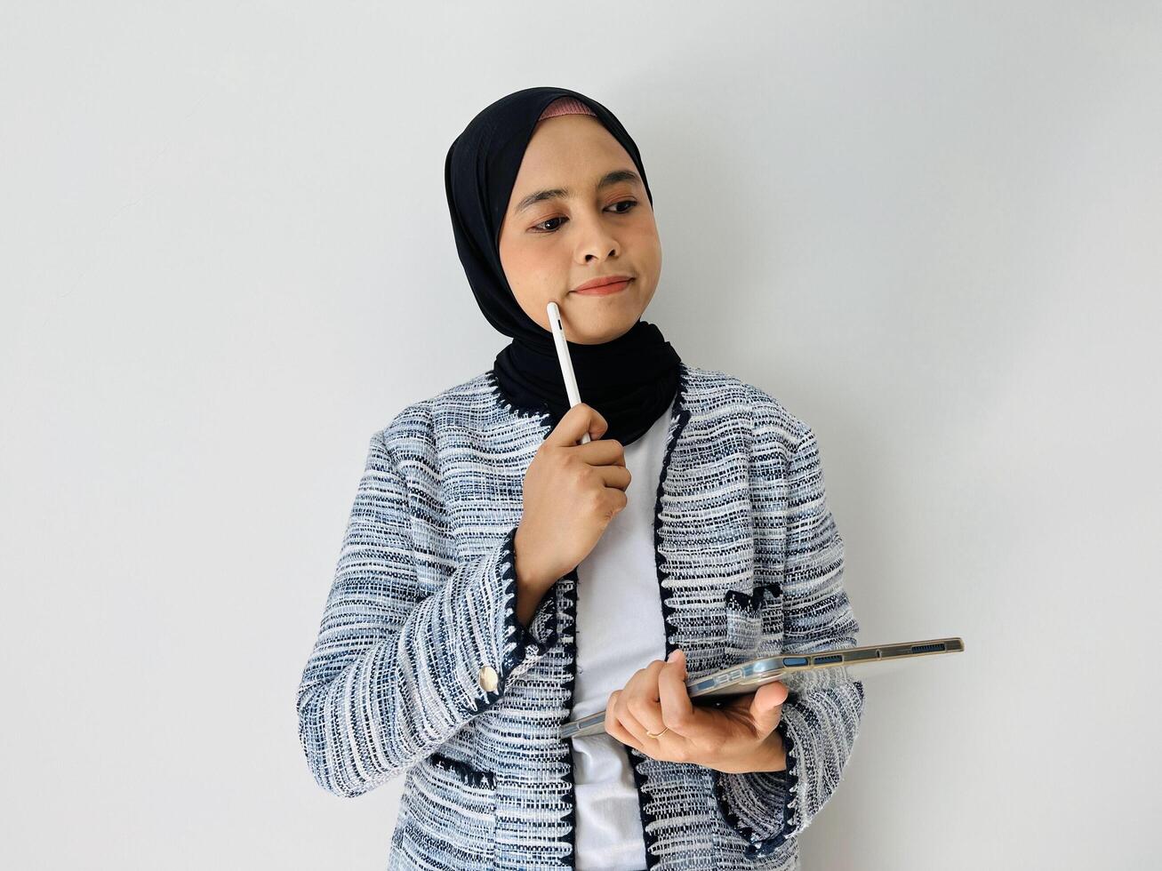A young Asian woman wearing black hijab and tweed blazer thinks deeply about something, holding digital tablet computer and keeps the pen near lips isolated by white background photo